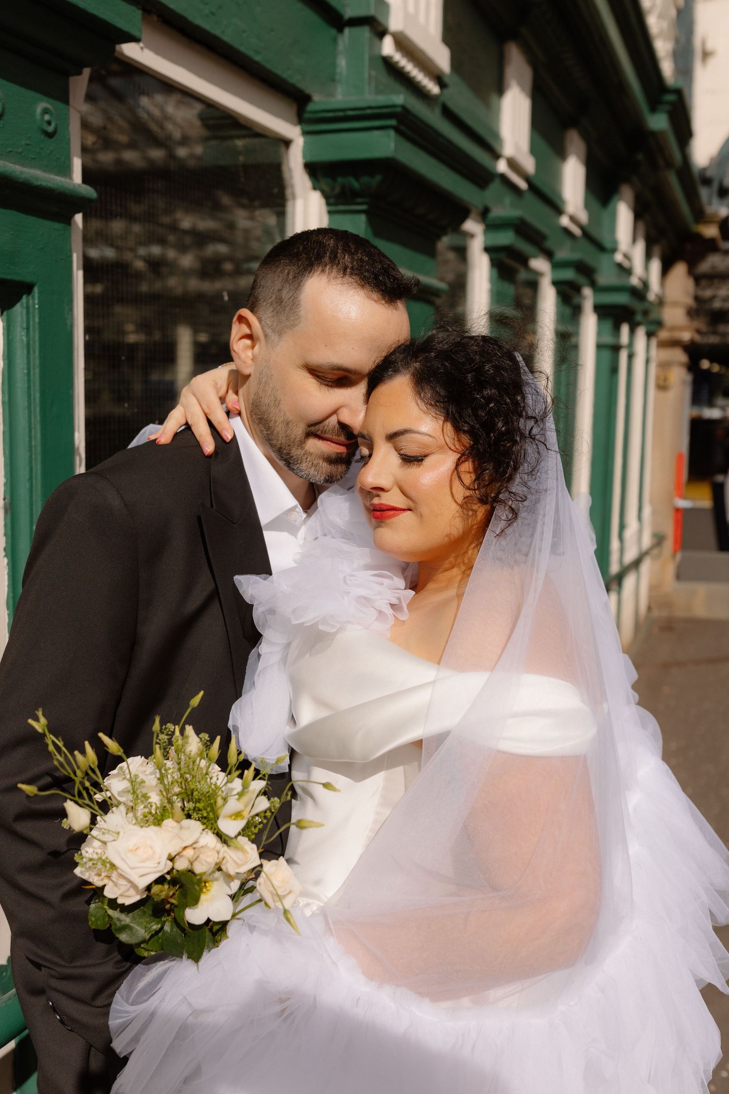 Bride and groom embrace outside green building, bride holds white bouquet wearing veil and white dress.