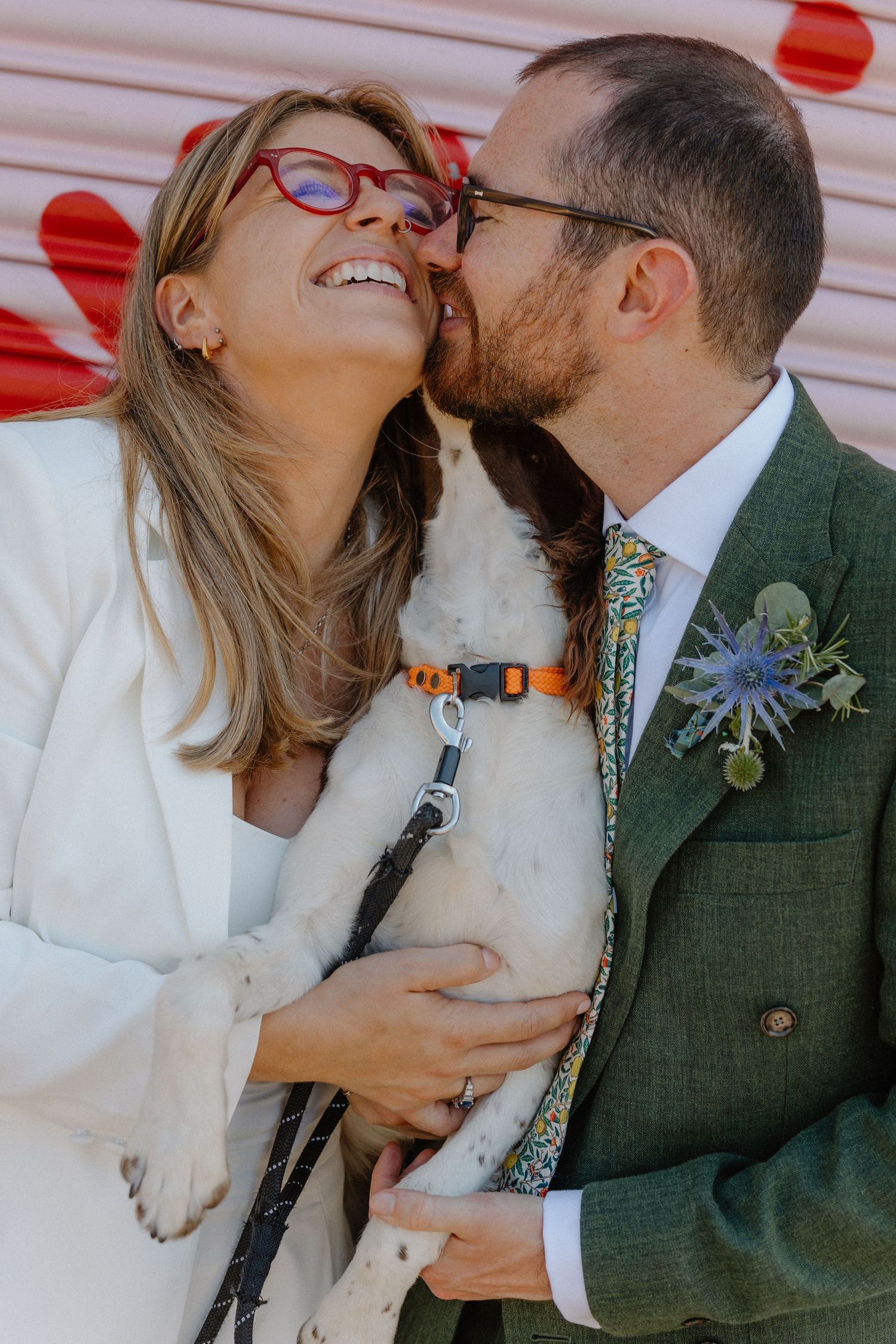 Groom in green suit kissing bride on the cheek while they hold their white spaniel dog outdoors.