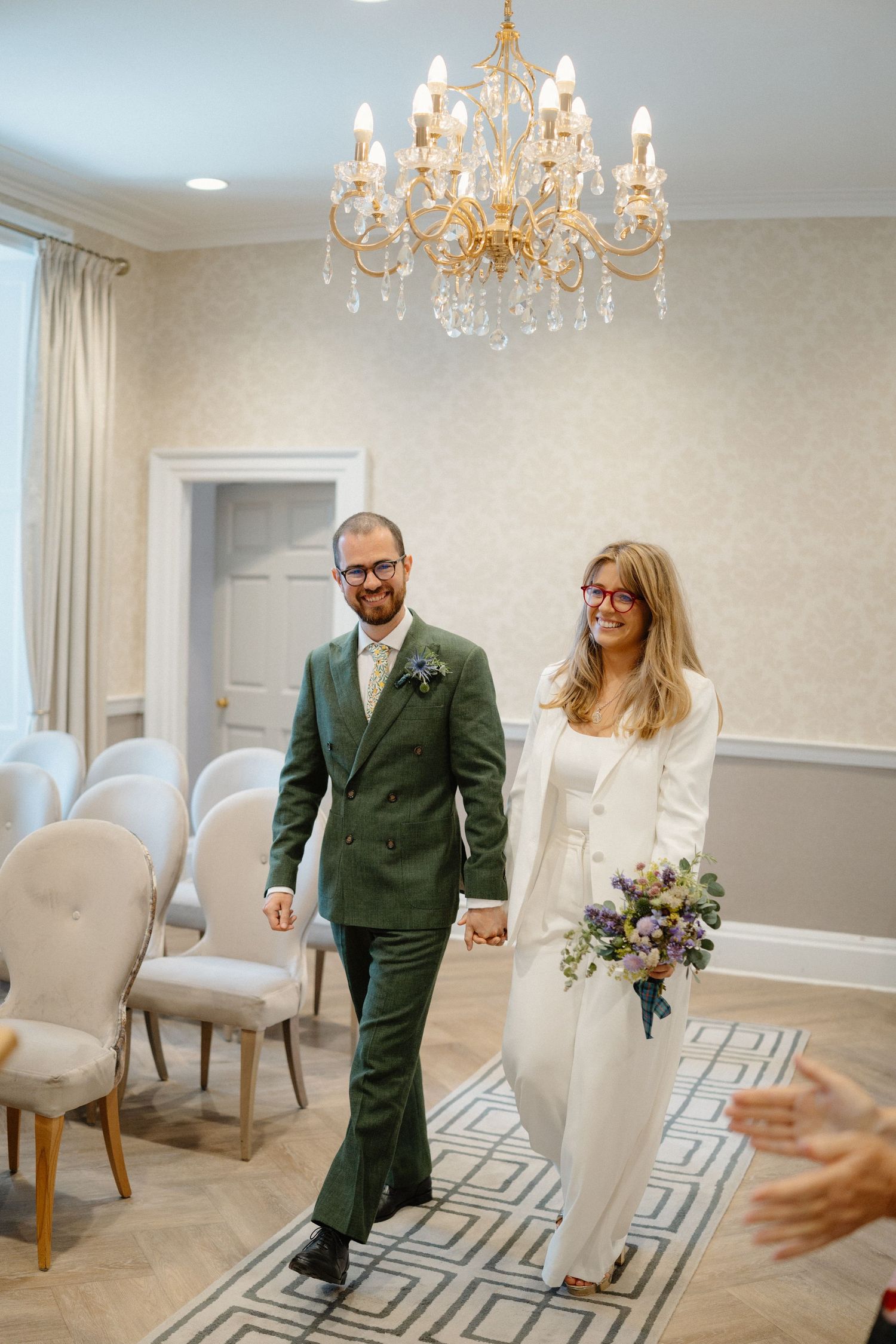 Newlyweds walk hand in hand beneath chandelier in elegant white ceremony room after exchanging vows.