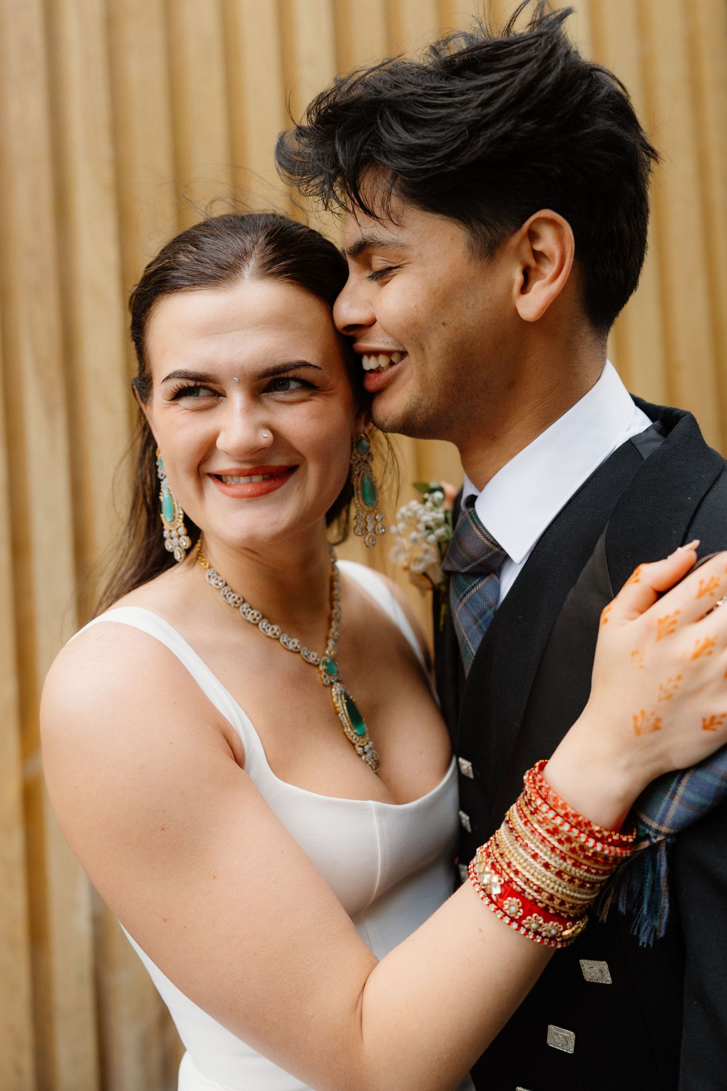 Bride and groom embrace smiling, she wears white dress with turquoise jewelry and henna bracelets, he wears dark suit with tie.