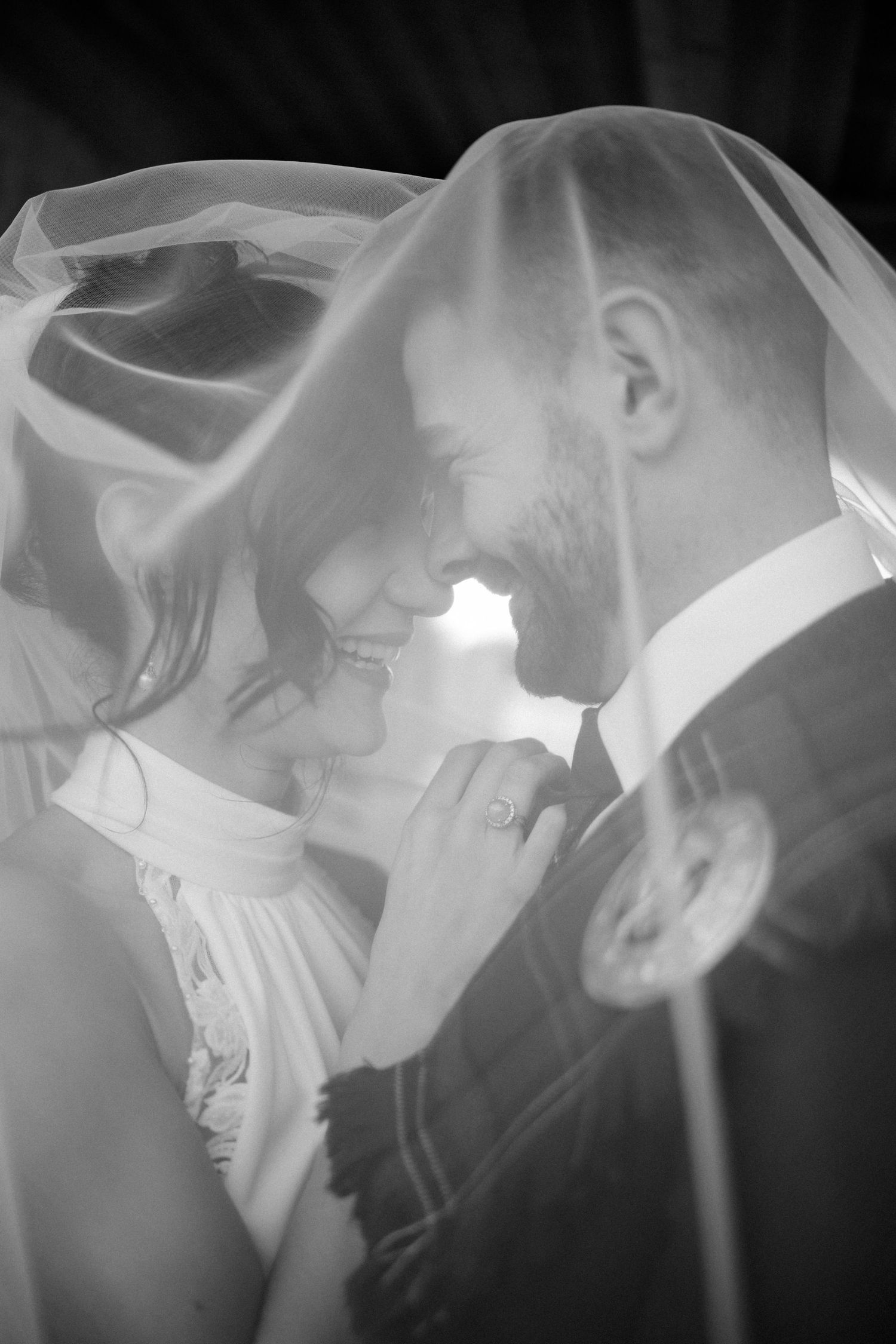 Black and white photo of bride and groom smiling under a wedding veil in an intimate moment.