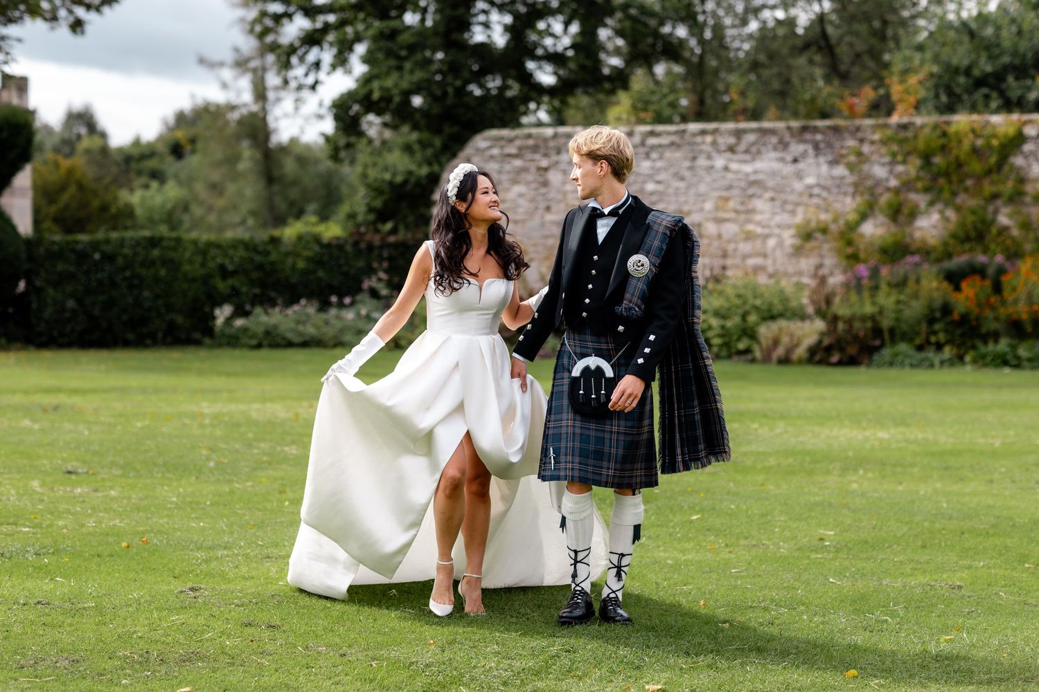 Bride in white gown and groom in blue tartan kilt walk hand in hand on green lawn at outdoor wedding.