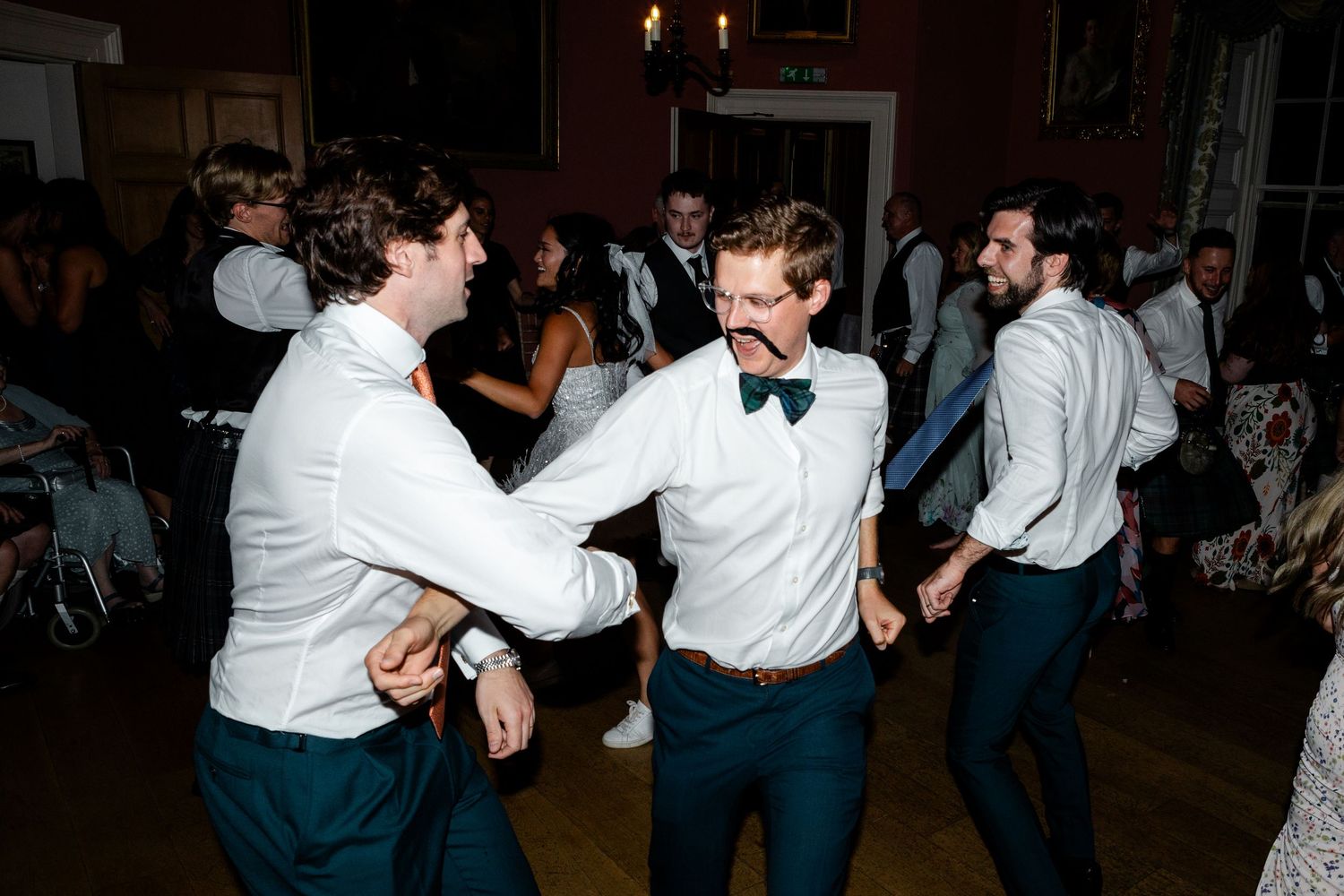 Energetic wedding guests in white shirts and bow ties dancing enthusiastically on the reception floor.