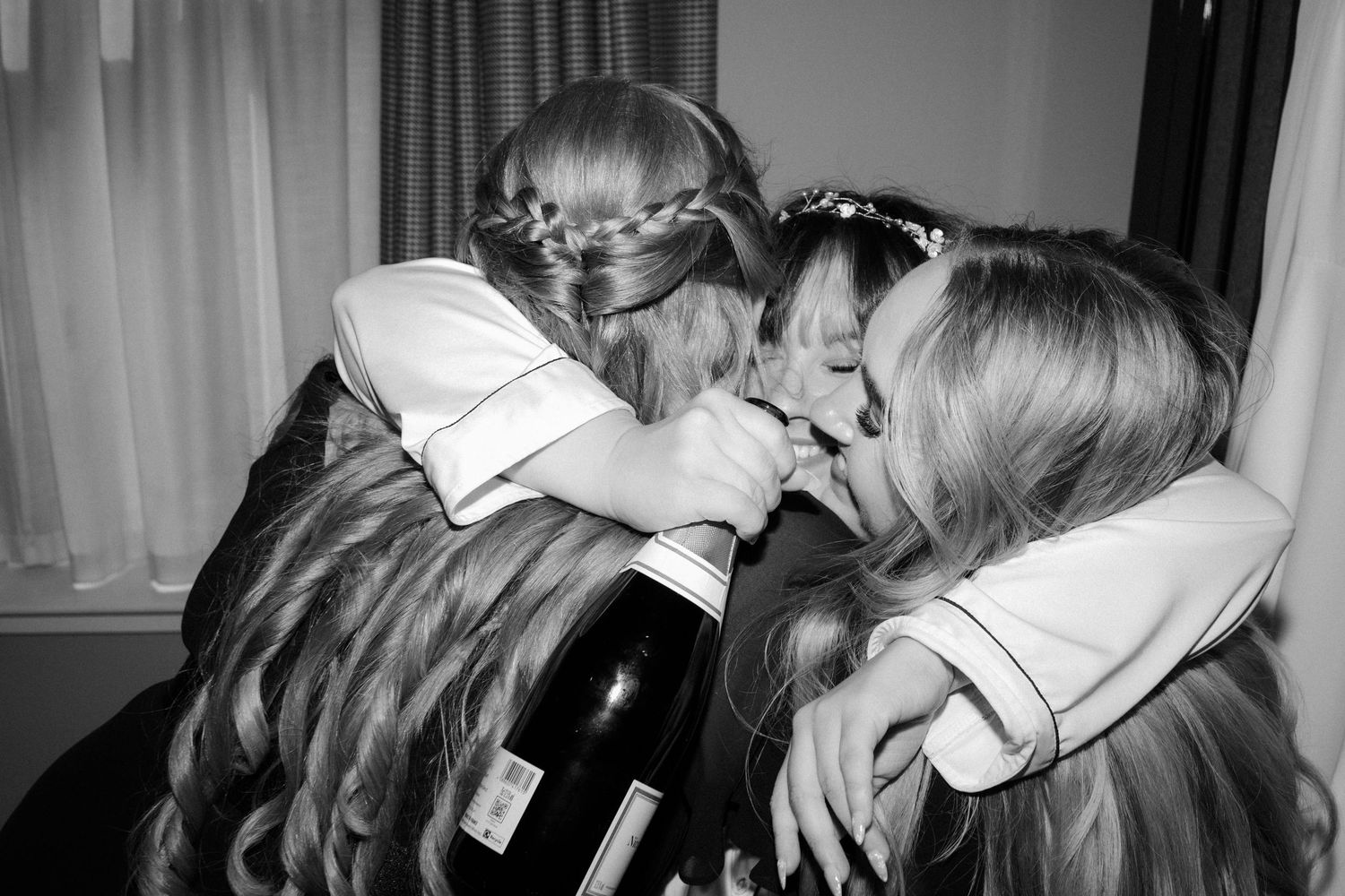 Black and white photo of bridesmaids hugging and sharing champagne bottle at a wedding celebration.