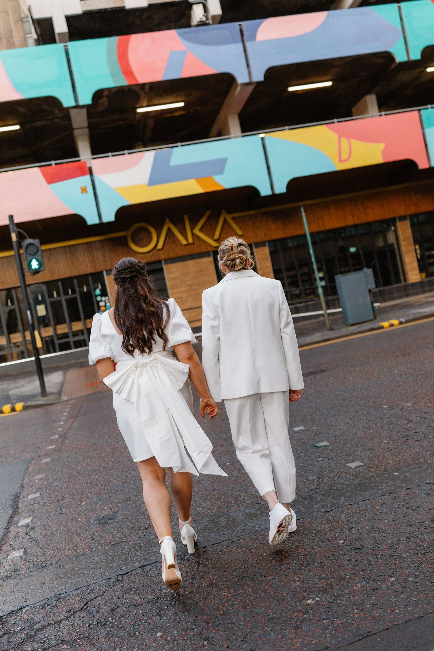 Couple in white outfits holding hands walking toward colorful OAKA building mural on city street.