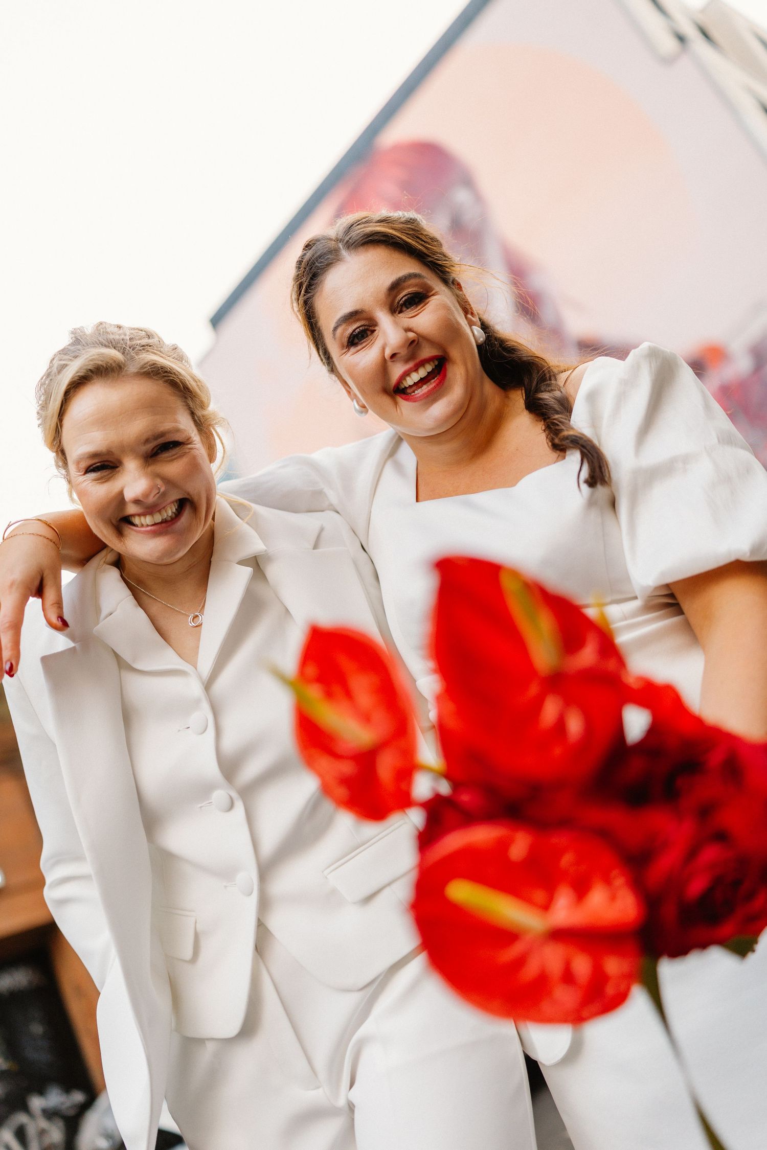 Two joyful brides in white smile together holding a bouquet of red anthurium flowers.