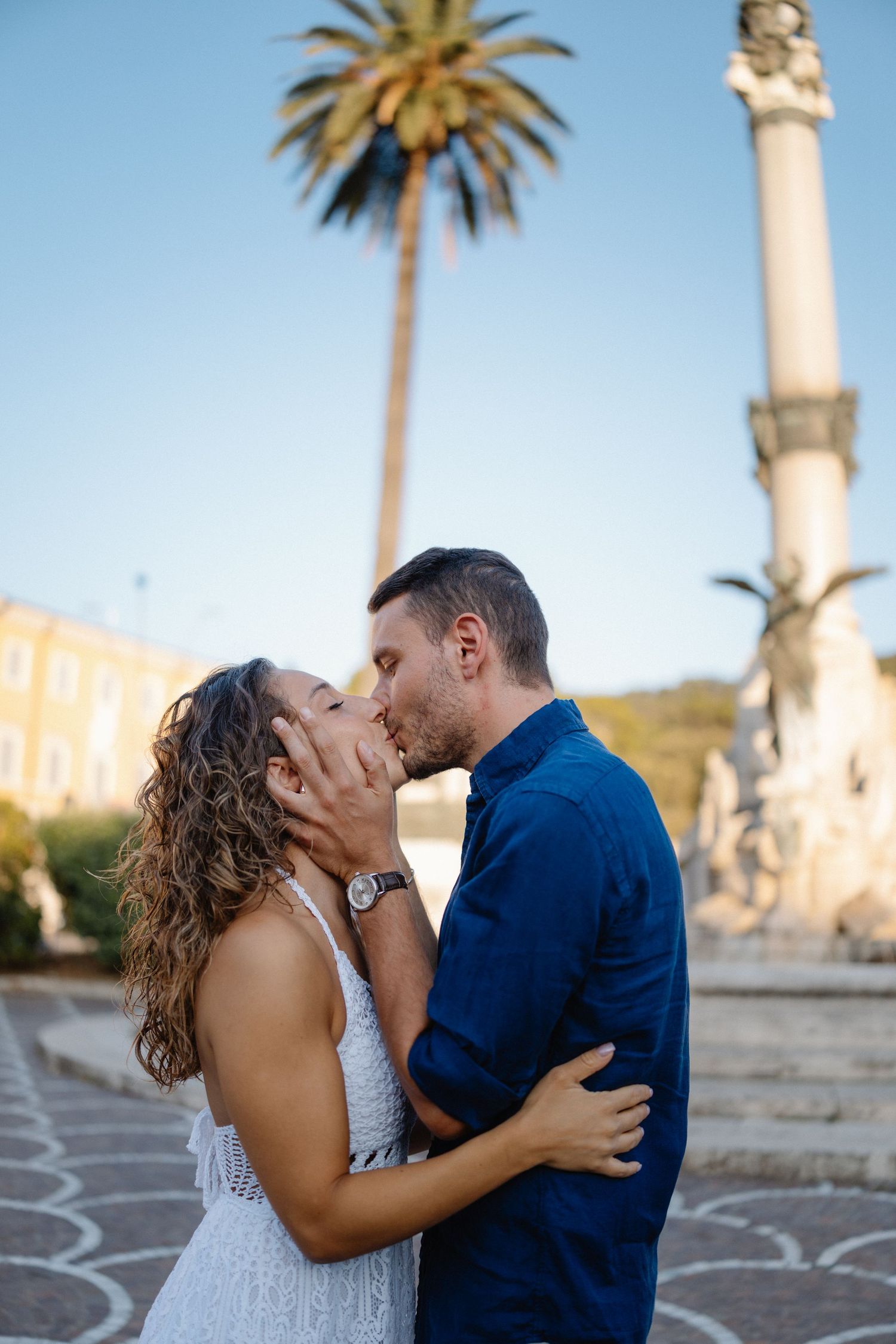 A couple shares an intimate moment at sunset by Mediterranean architecture with palm trees.