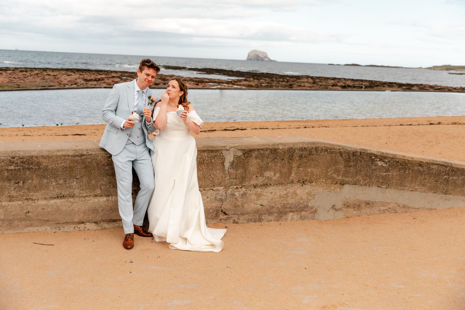 Couple sitting together on coastal rocks with ocean and beach in background.