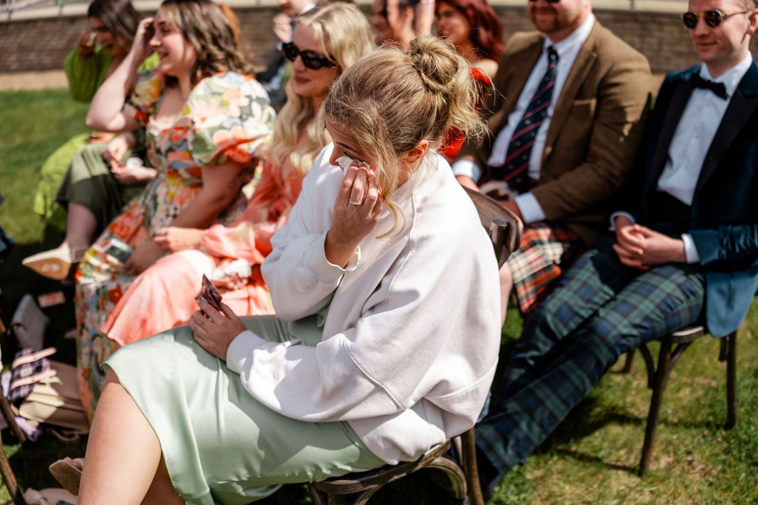 Wedding guests laugh and react emotionally during an outdoor ceremony on a sunny day.