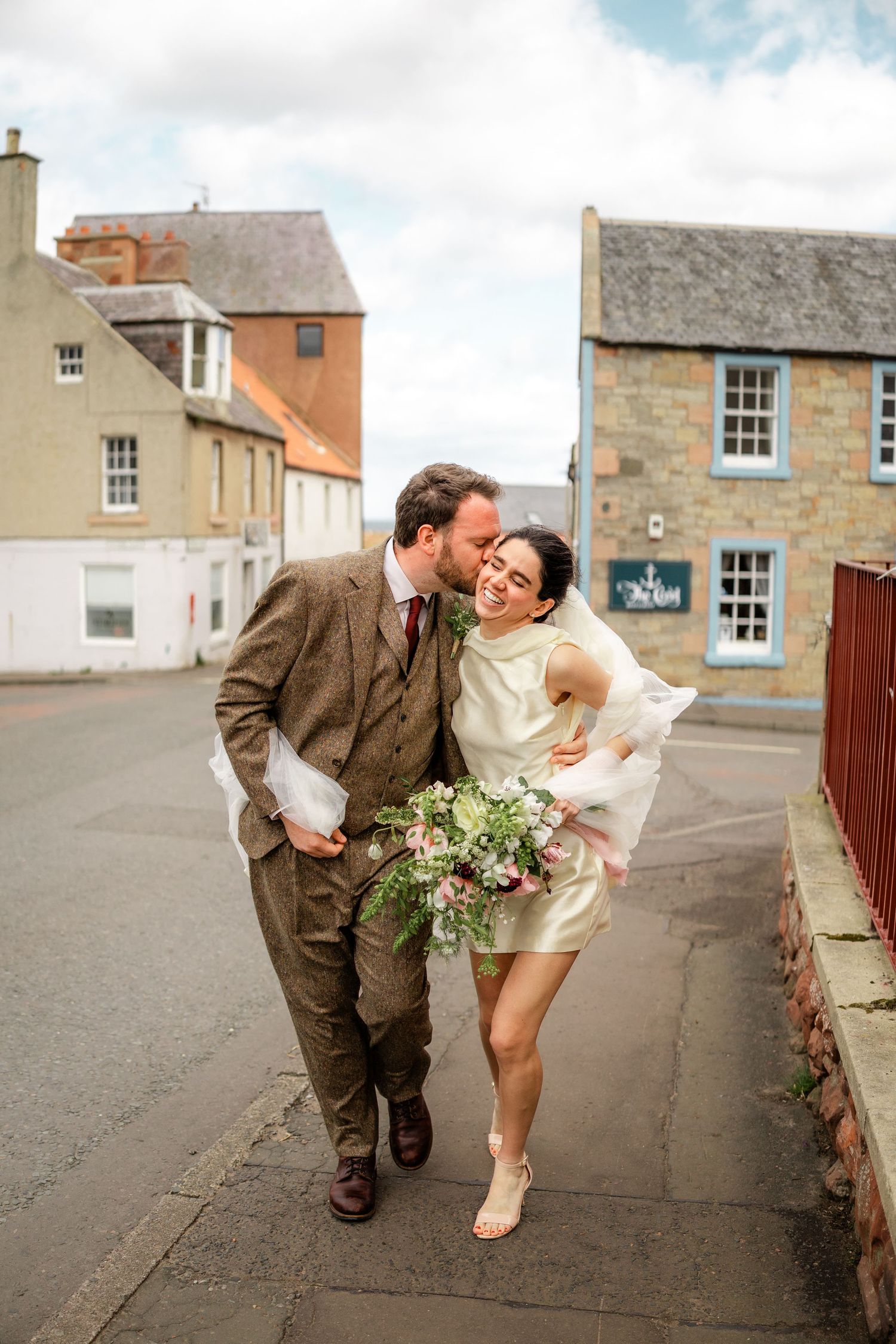 Wedding couple shares a playful moment on a Scottish village street with stone buildings.