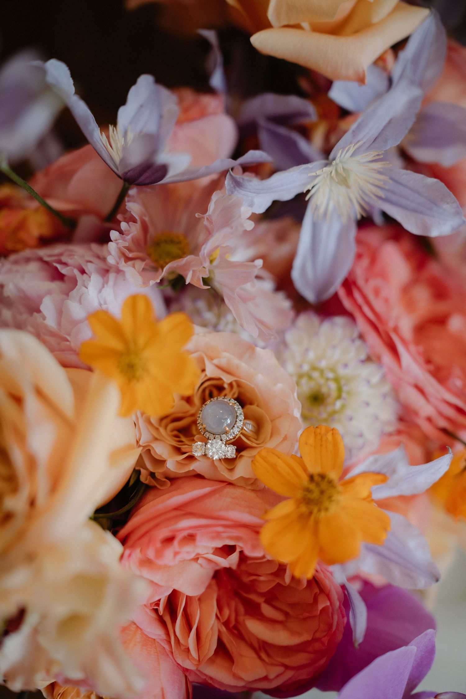 A diamond engagement ring nestled among pink, orange and white flowers in a romantic floral arrangement.