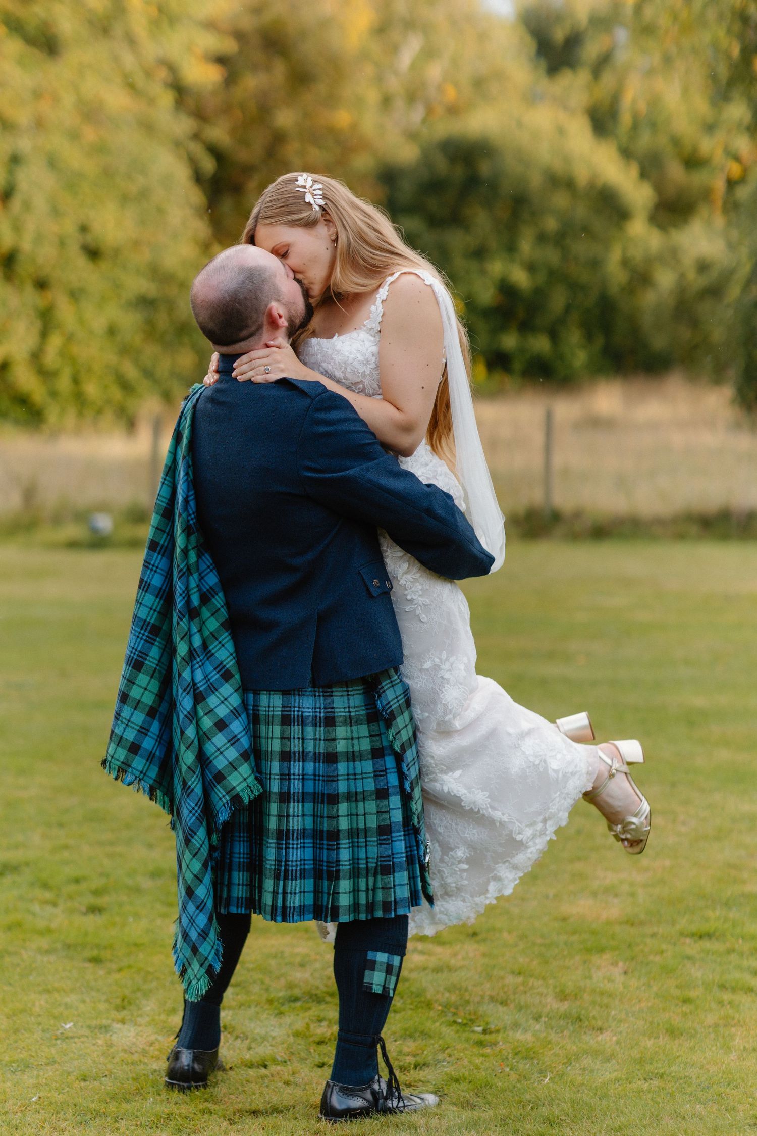 A couple in wedding attire dances together on green grass with the groom wearing a traditional Scottish kilt.
