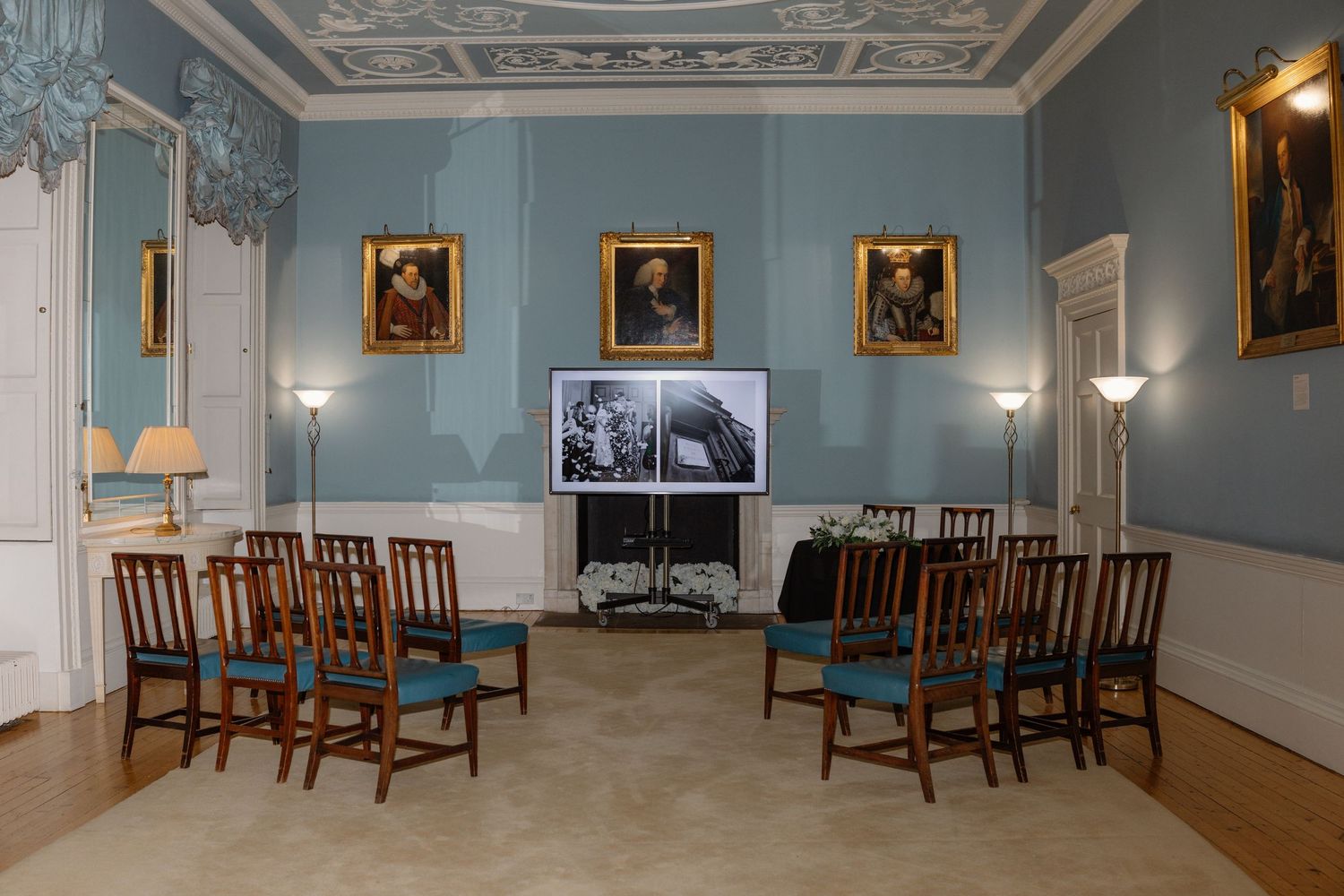 Elegant blue-walled room with ornate ceiling, antique furniture, and framed portraits displayed in a historic mansion interior.