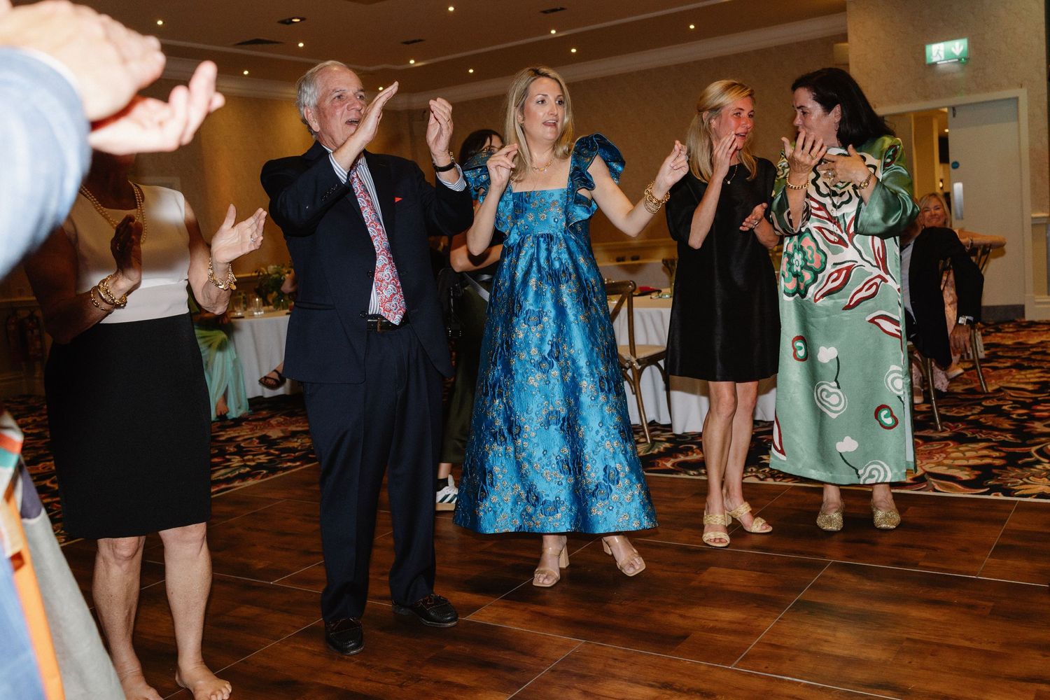 Wedding guests in formal attire dancing and clapping joyfully on dance floor at reception celebration.