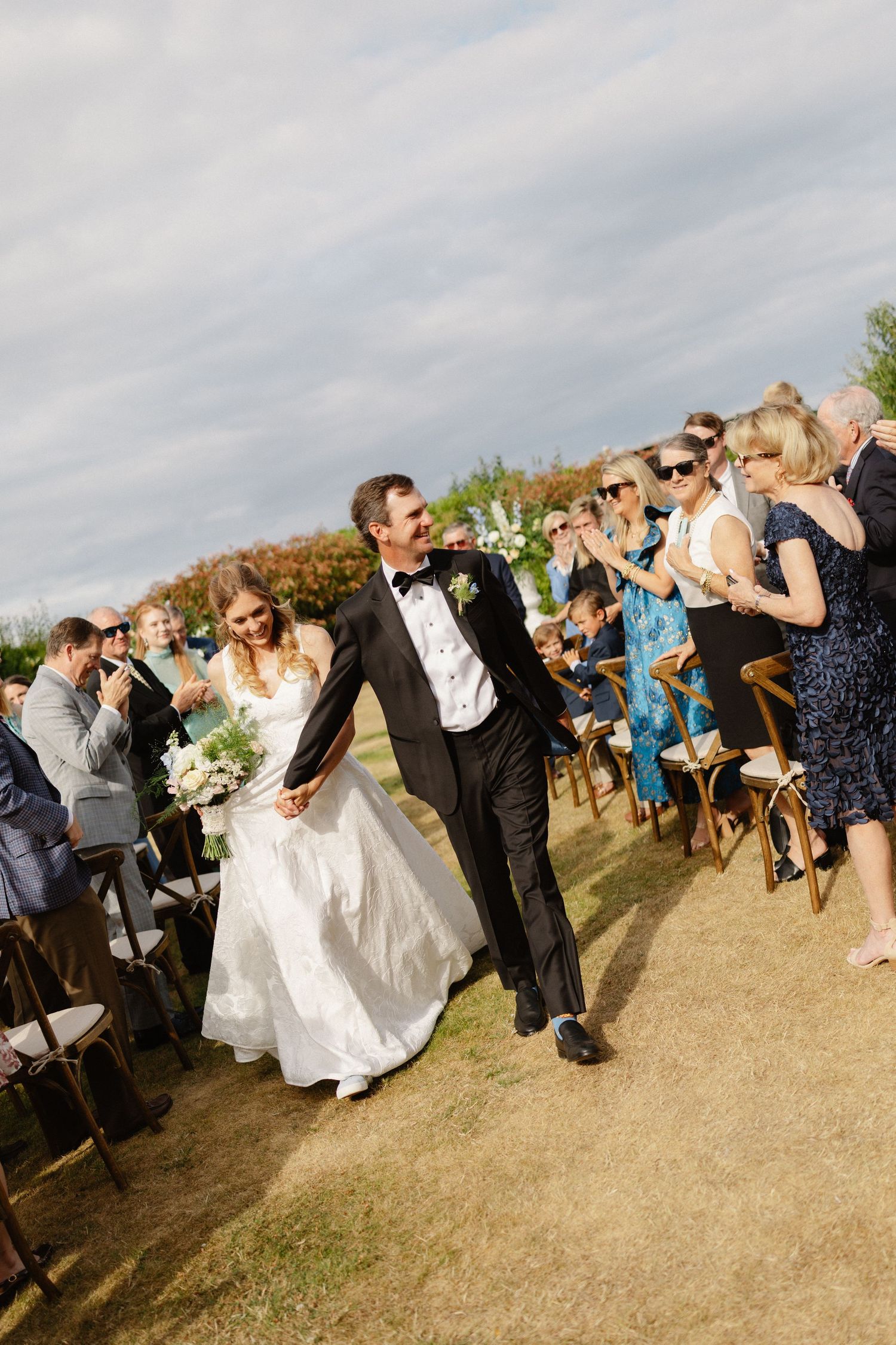 Joyful newlyweds walk down aisle after outdoor wedding ceremony as guests celebrate.