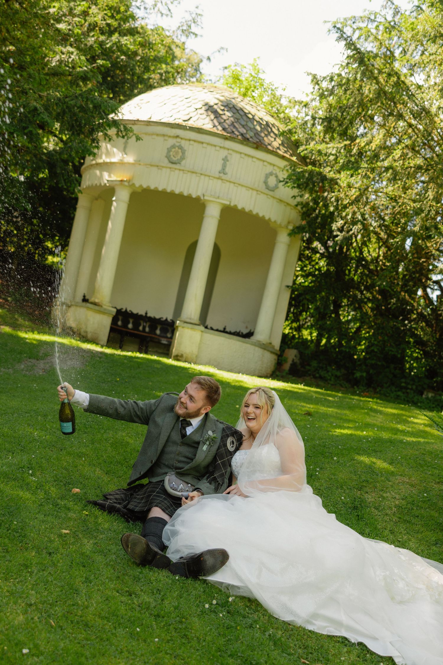 Bride and groom sitting on grass popping champagne in front of a white gazebo on their wedding day.