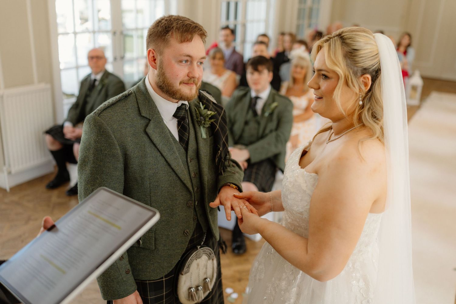 Bride placing wedding ring on grooms finger during Scottish wedding ceremony in kilt.