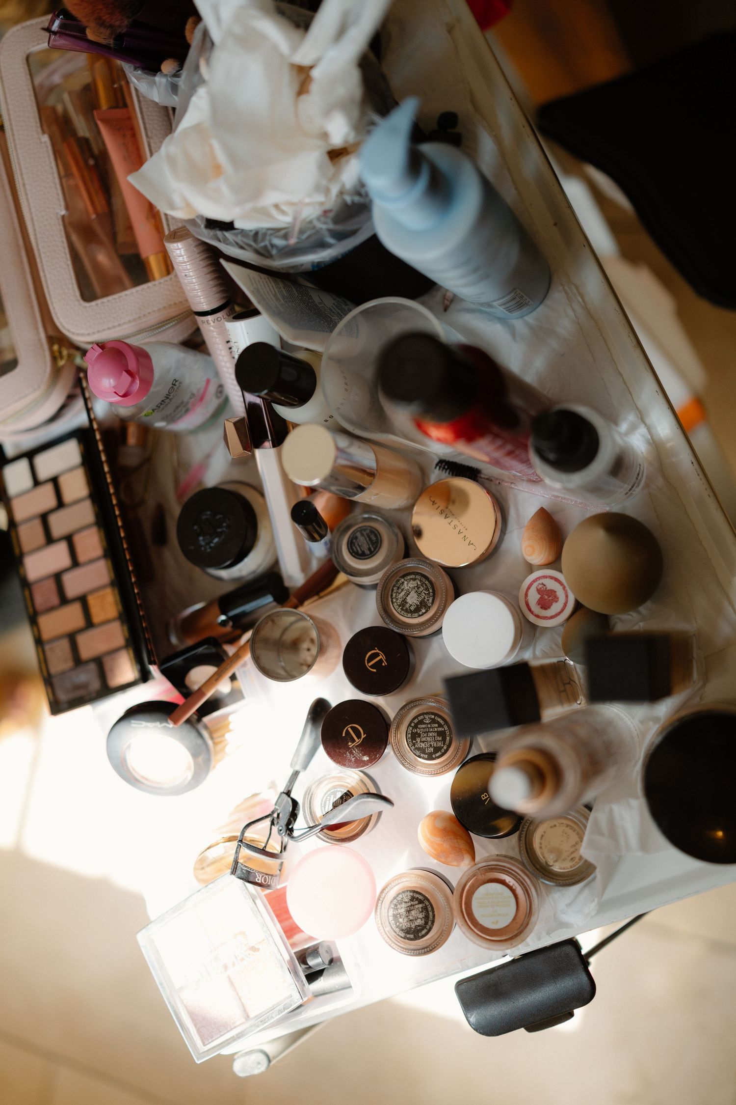 Overhead view of a cluttered makeup vanity table covered with various cosmetics, brushes, and beauty products.