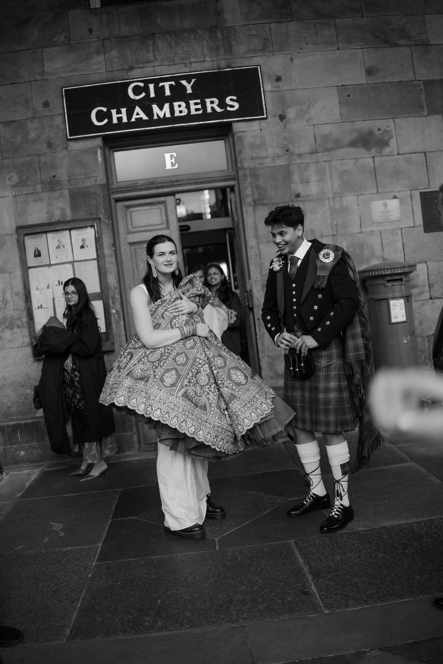 Bride holding embroidered lehenga and groom in Scottish kilt outside Edinburgh City Chambers in black and white.