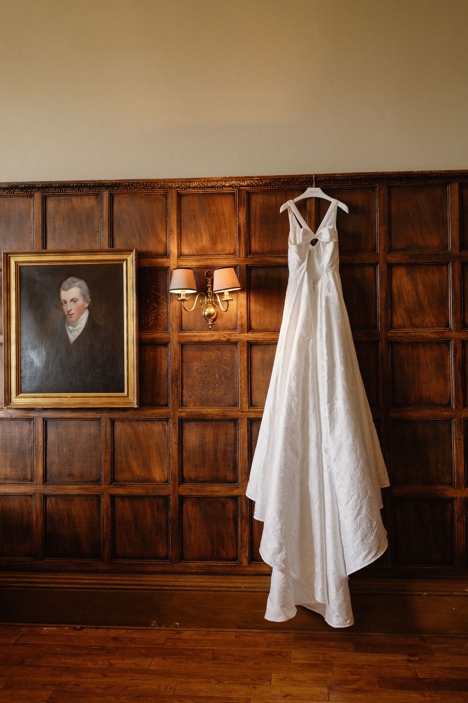 White wedding gown hanging on wooden paneled wall beside vintage portrait painting and wall sconce.