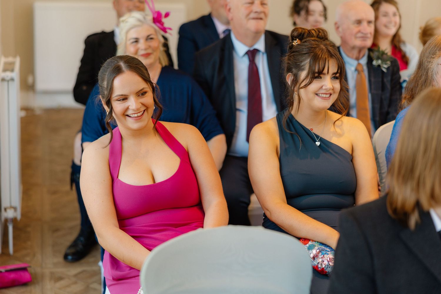 Two bridesmaids smiling and laughing during a wedding ceremony, one in pink and one in navy blue dress.