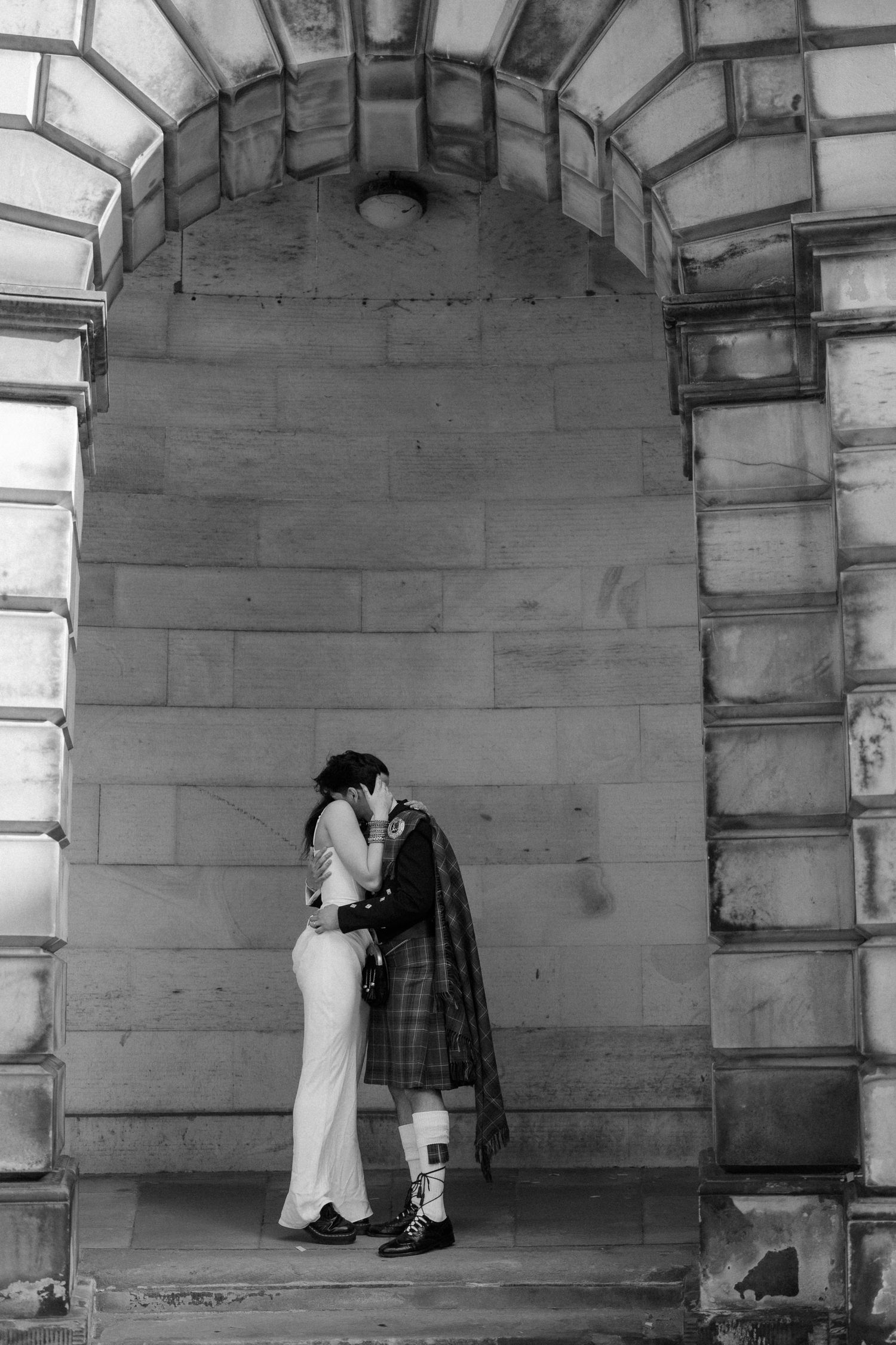 Black and white photo of a couple kissing under a stone archway, groom in Scottish kilt, bride in elegant white dress.