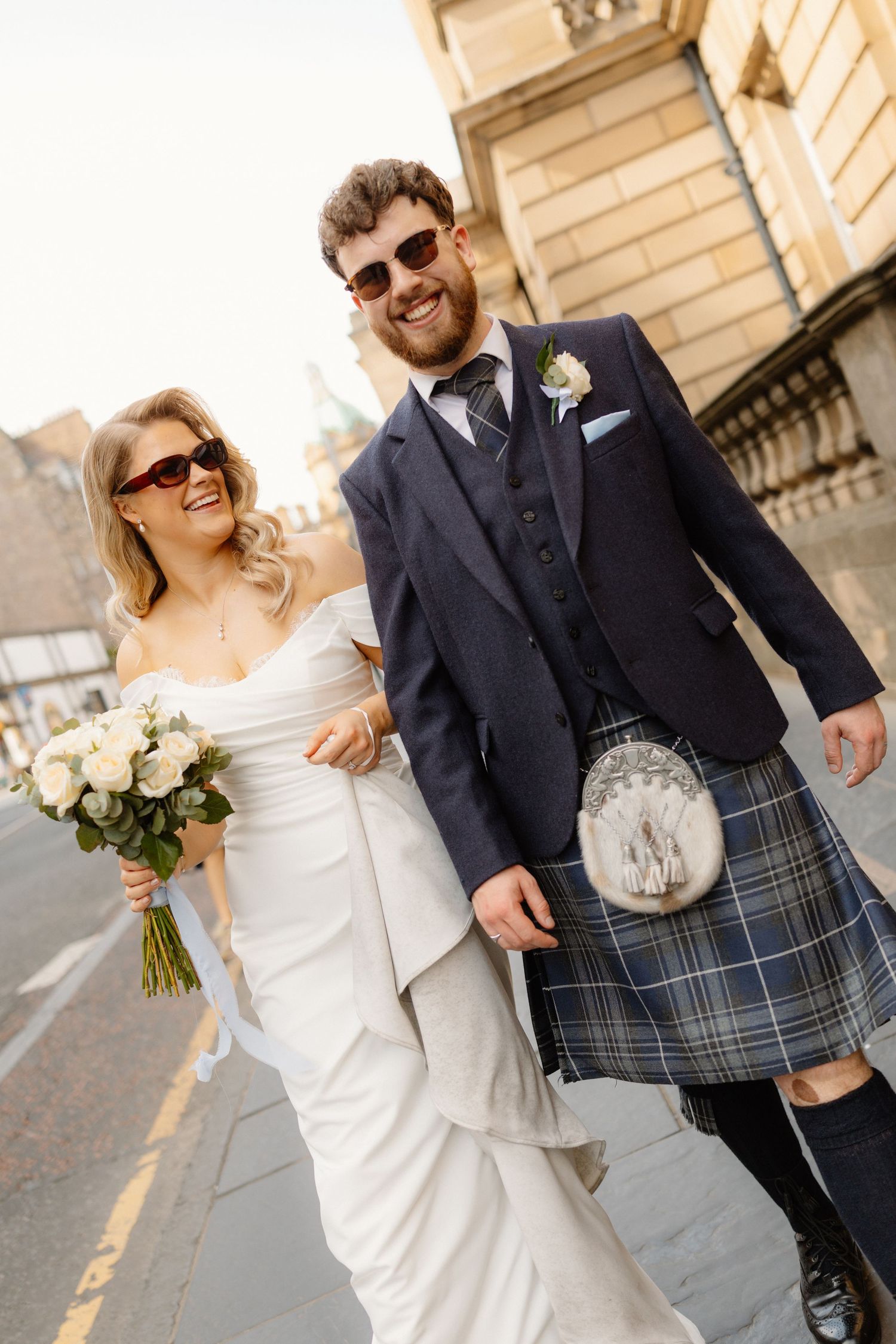 Smiling bride and groom in Scottish kilt walking outdoors on wedding day in Edinburgh city center.
