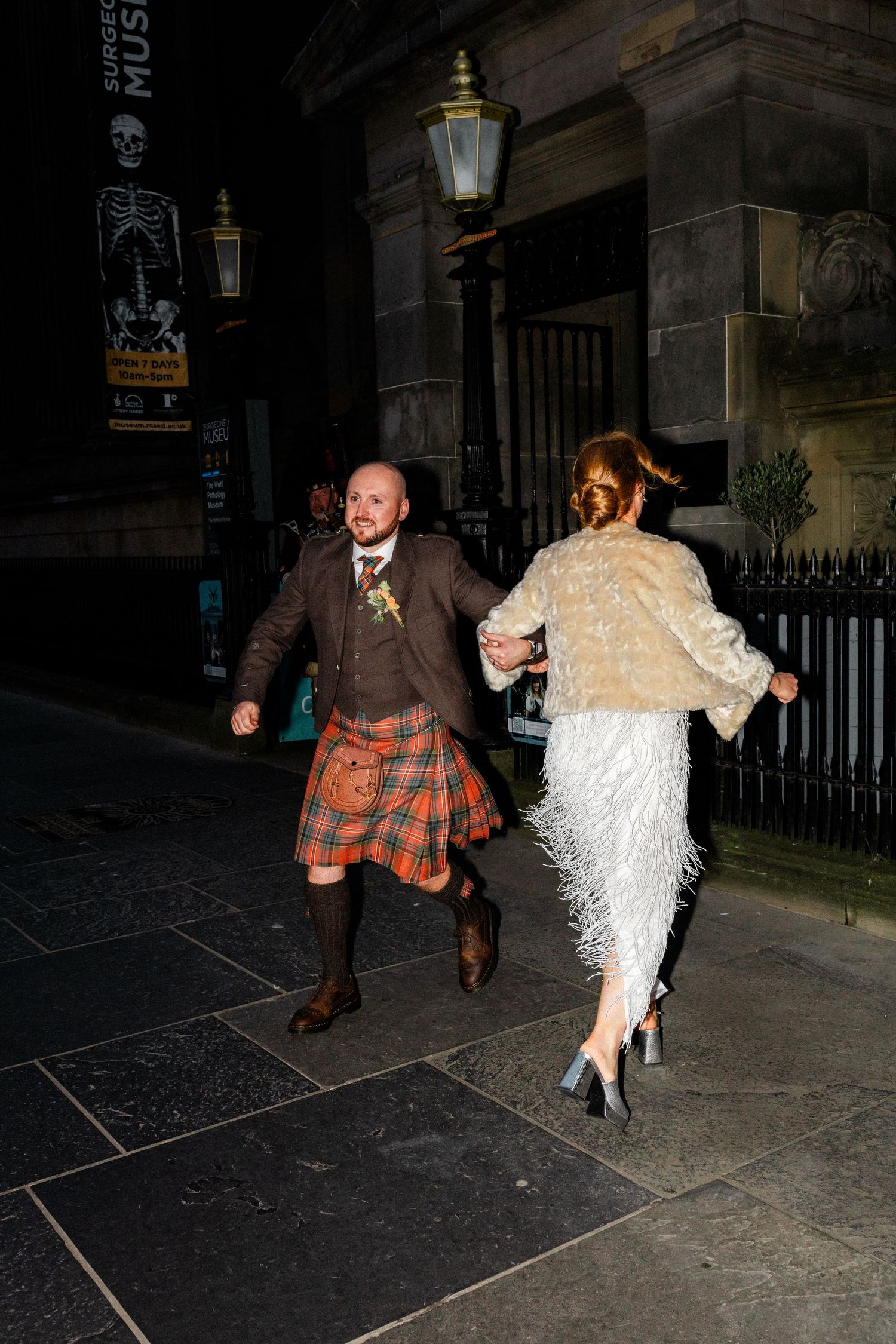 A couple in wedding attire walking at night, with the groom wearing a kilt and brown jacket.