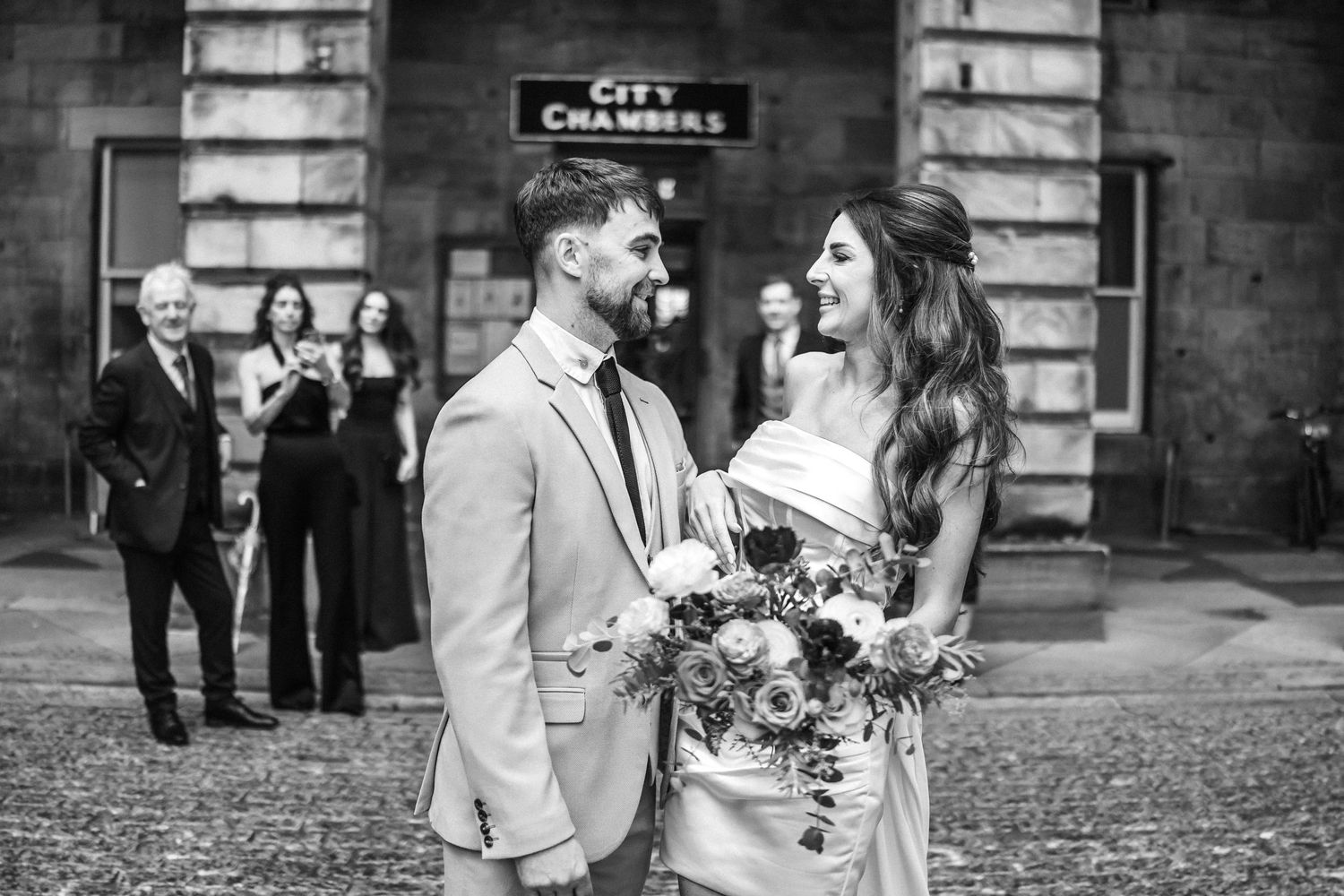 A black and white wedding photo showing a couple sharing a joyful moment outside a historic stone building.