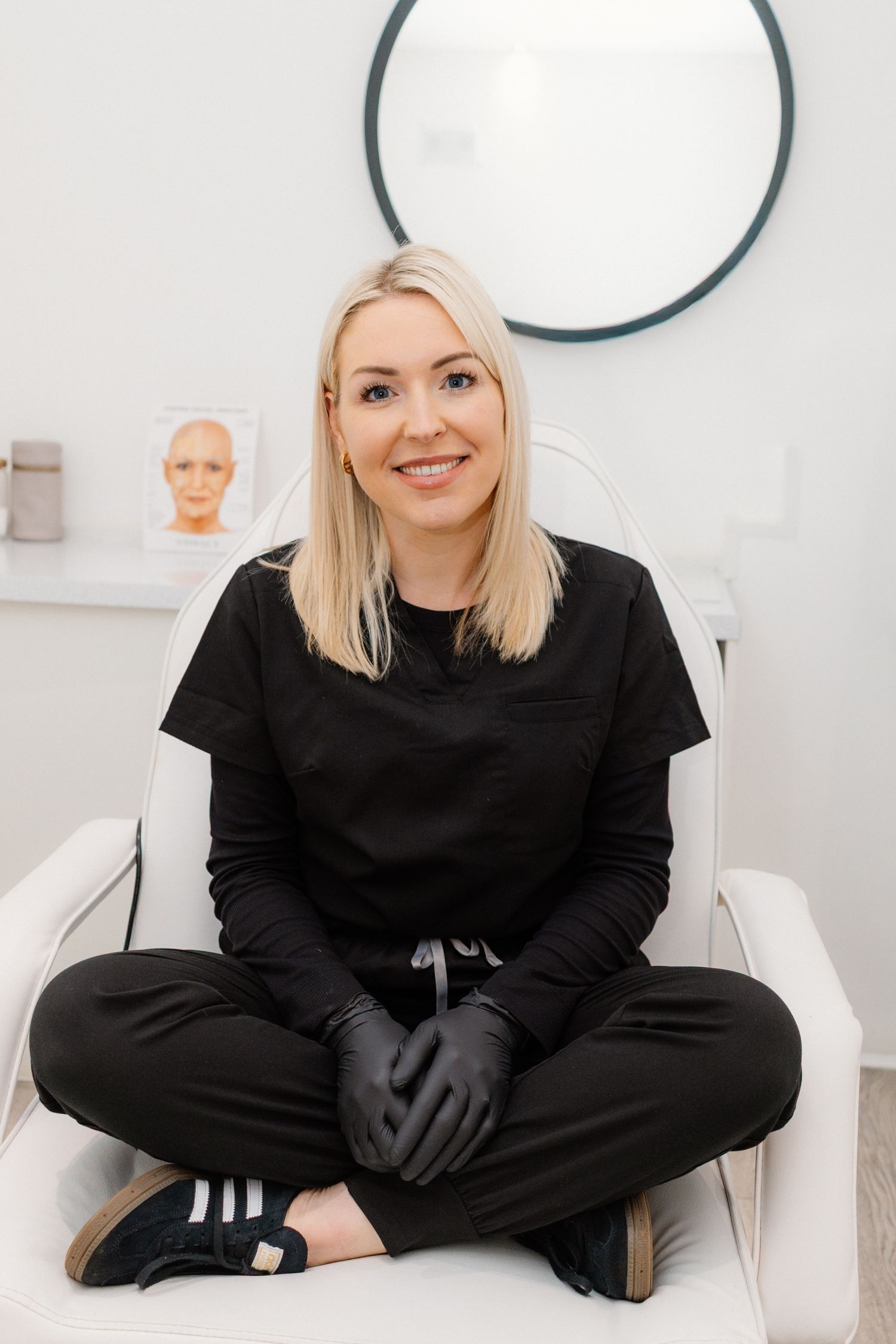 Healthcare professional in black outfit sitting cross-legged in medical office.