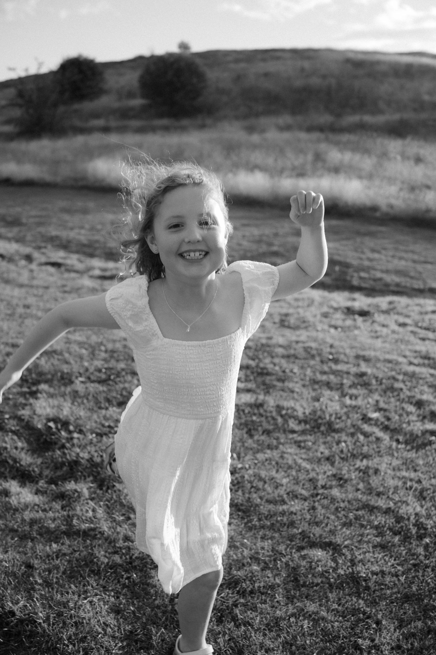 Child in white dress runs and plays in grassy field during black and white photoshoot.