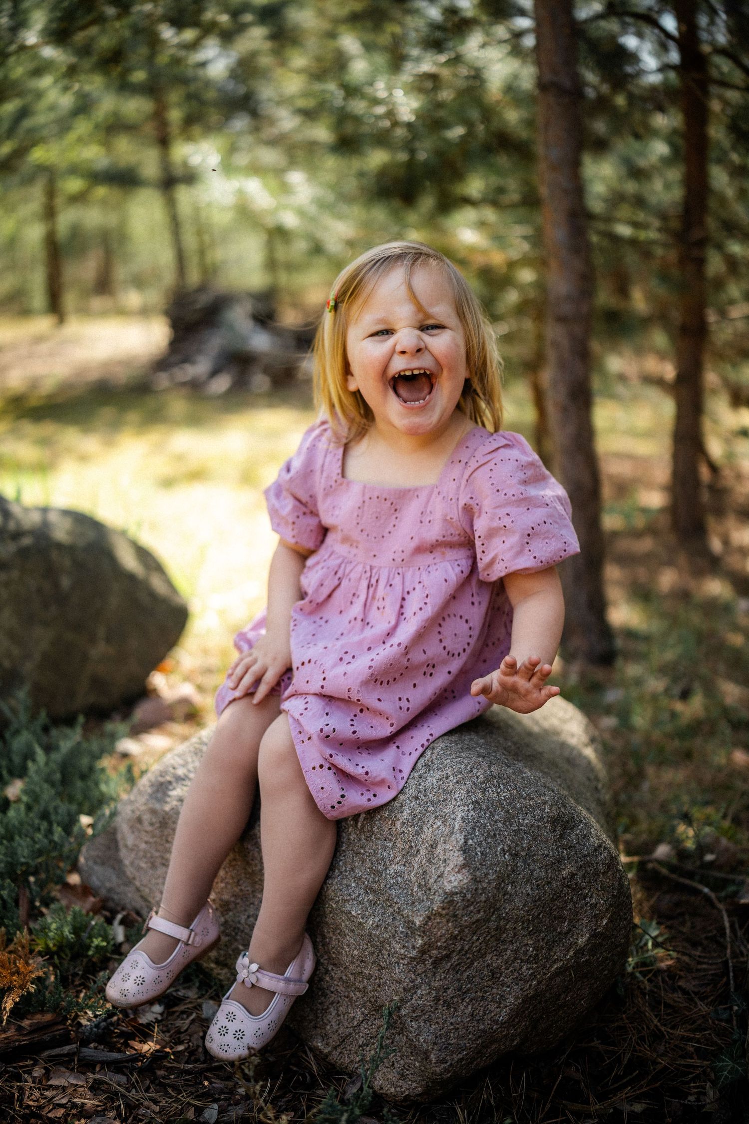 A child in a pink dress laughs joyfully while sitting on a large rock in a forest setting.