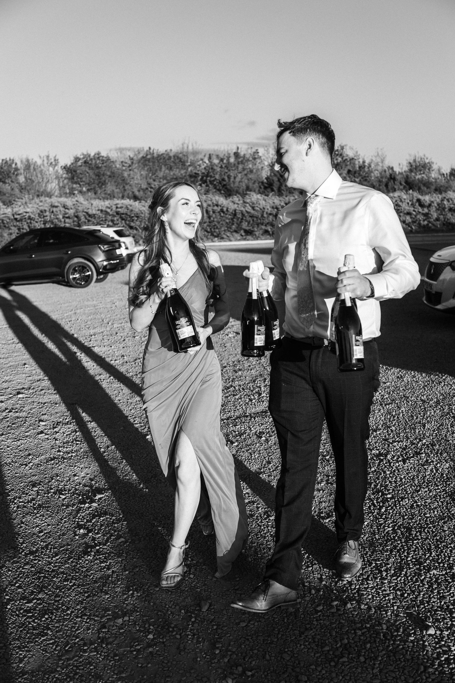 Black and white photo of an elegant couple holding wine bottles outdoors, smiling and conversing near parked cars.