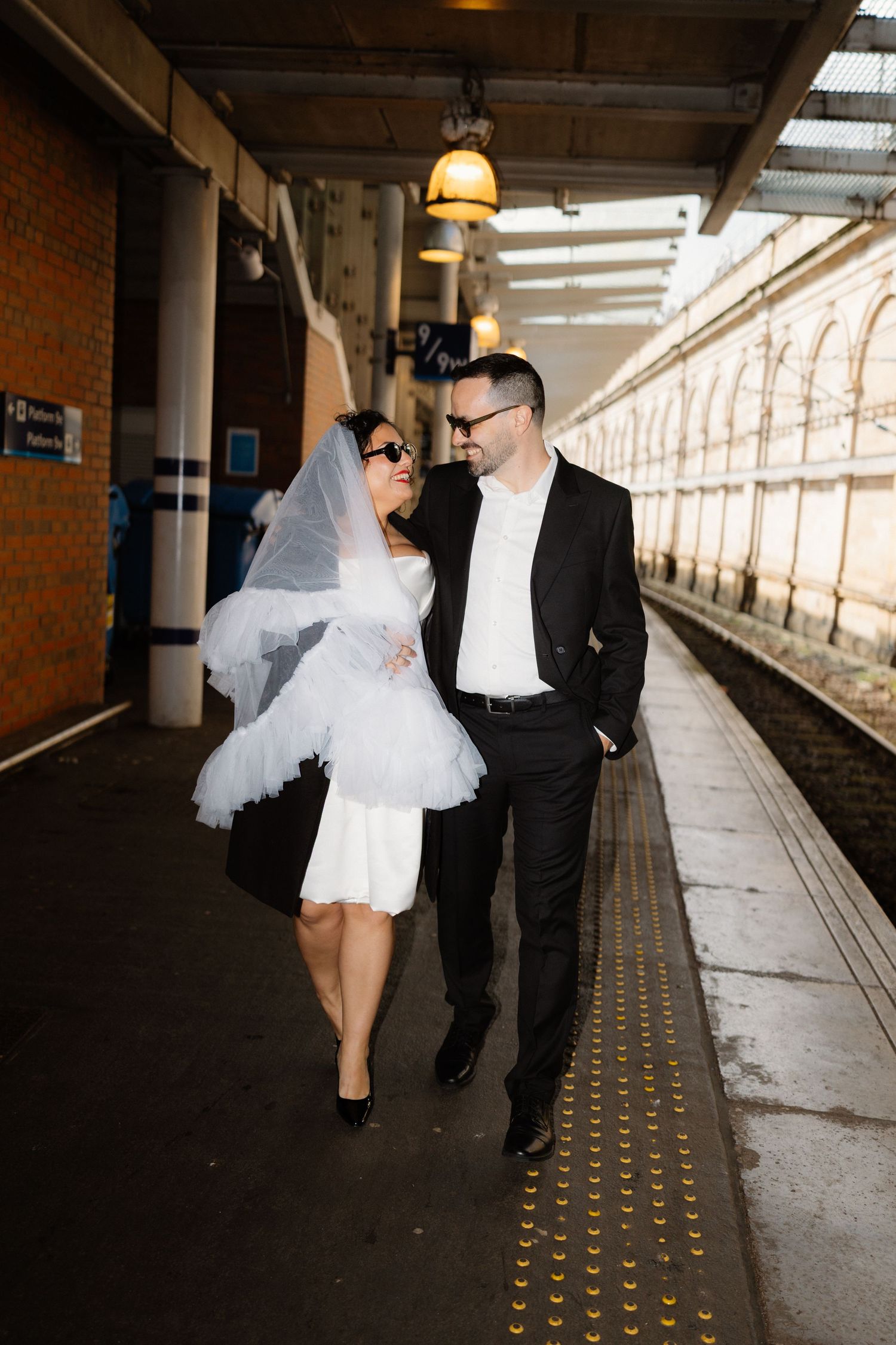 Bride and groom walking on a train platform, bride wearing short white dress and veil with sunglasses.