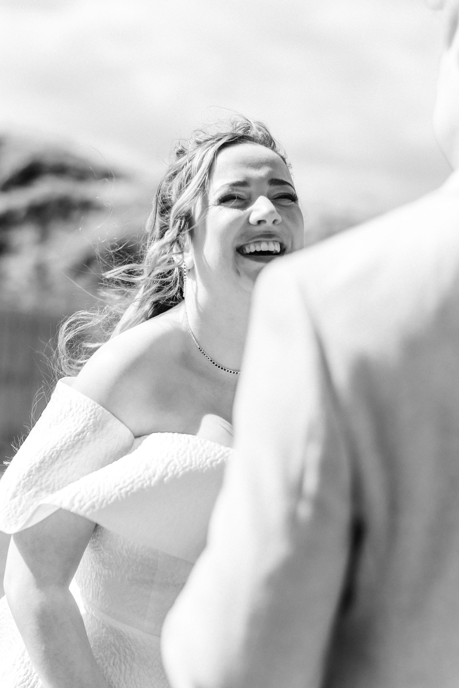Bride in off-shoulder white dress laughing joyfully outdoors on her wedding day.