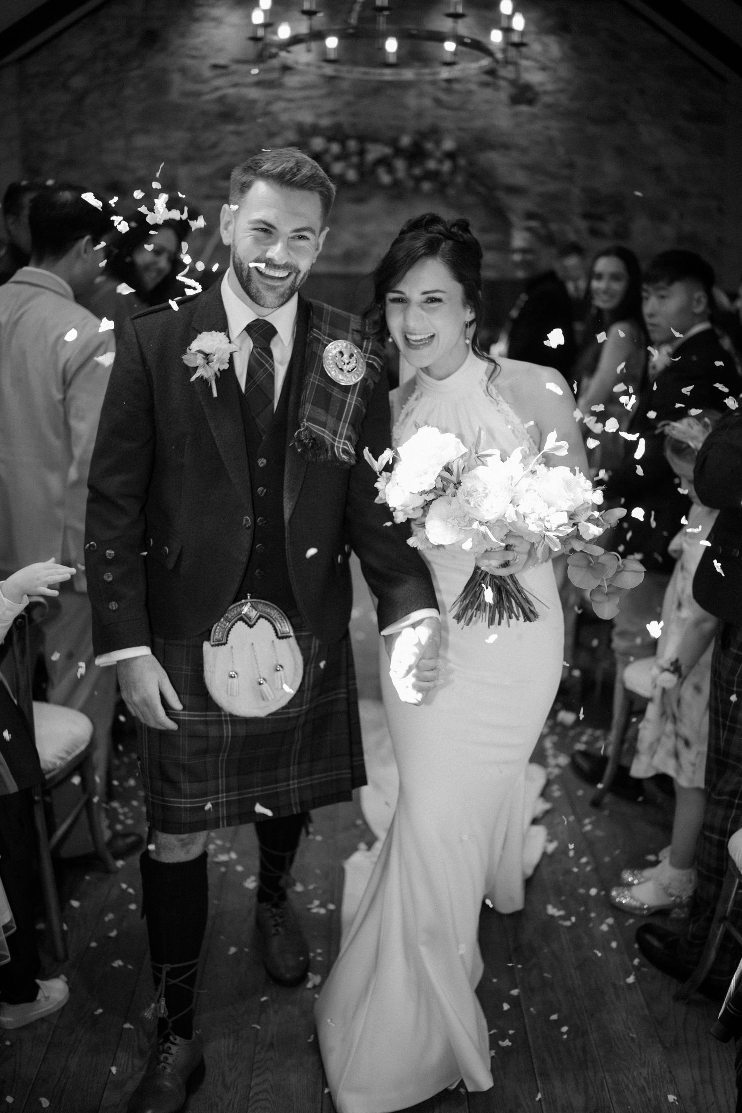 Black and white photo of newlyweds walking down the aisle as guests throw confetti, groom wearing Scottish kilt.