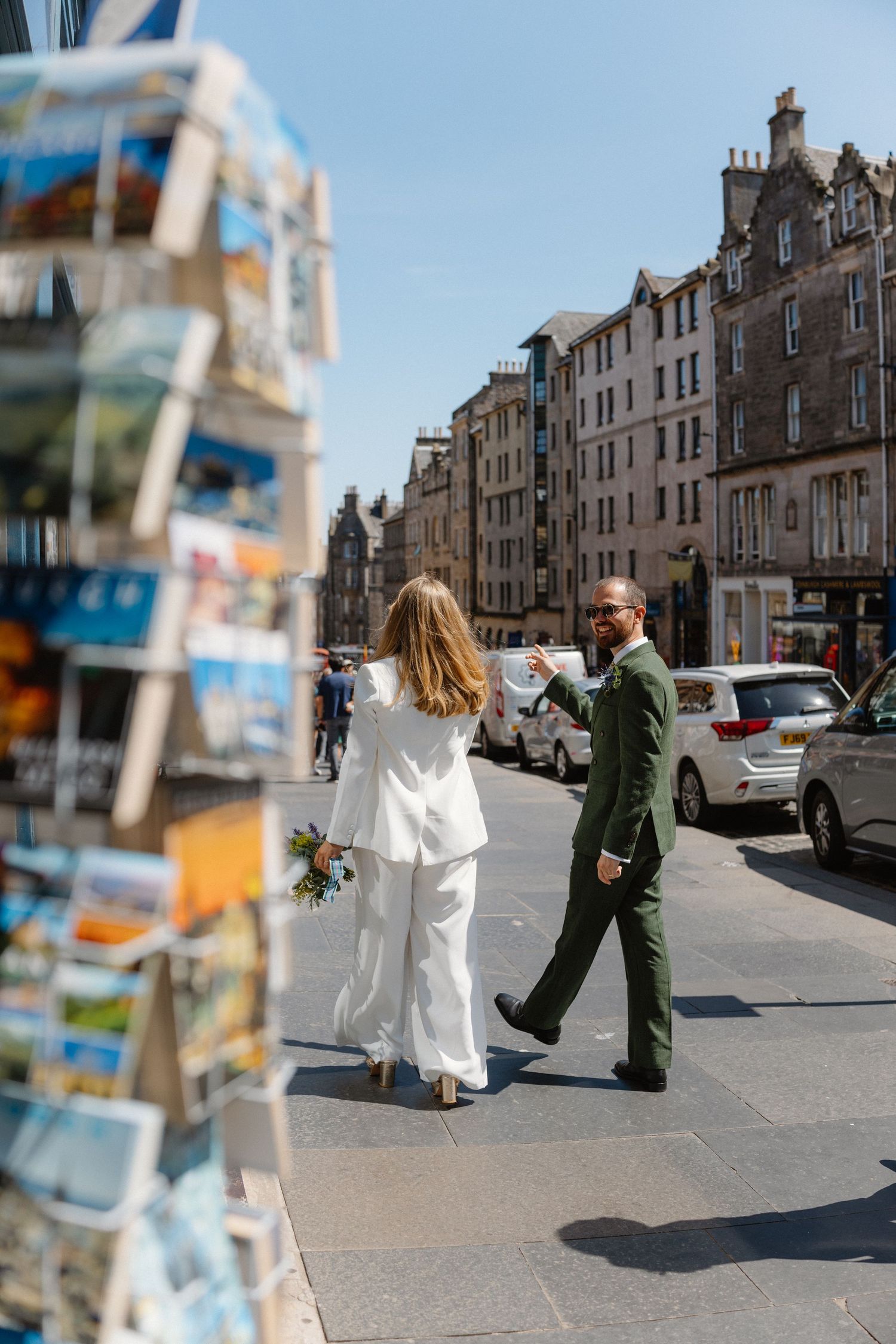 Bride and groom in wedding attire browsing postcard stand on sunny Edinburgh city street.