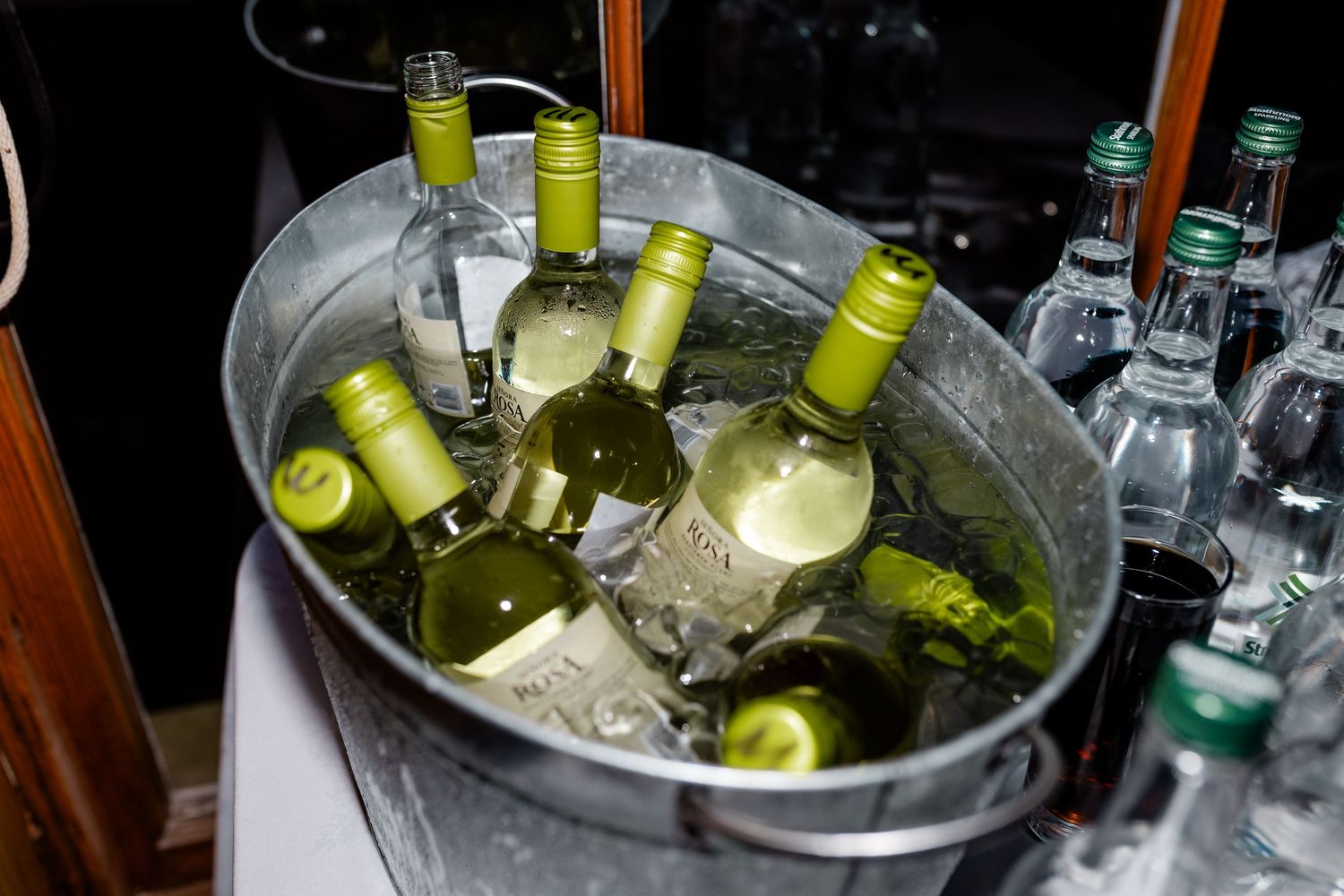 Chilled white wine and water bottles in an ice-filled galvanized metal bucket at a wedding bar.