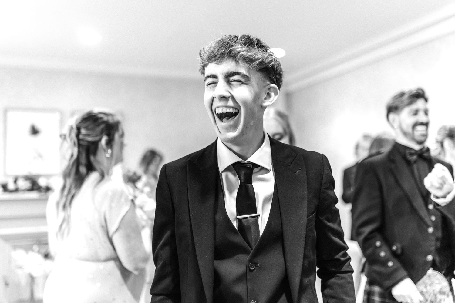 Young man in a suit laughing joyfully at a wedding celebration indoors.