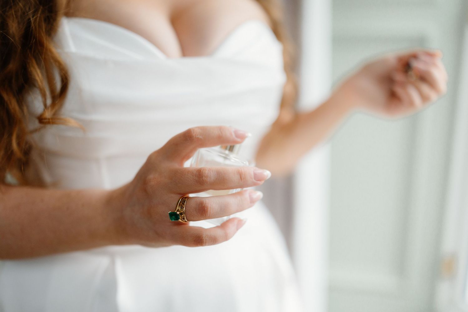 Bride in white strapless gown spraying perfume, wearing gold emerald ring, soft natural light.