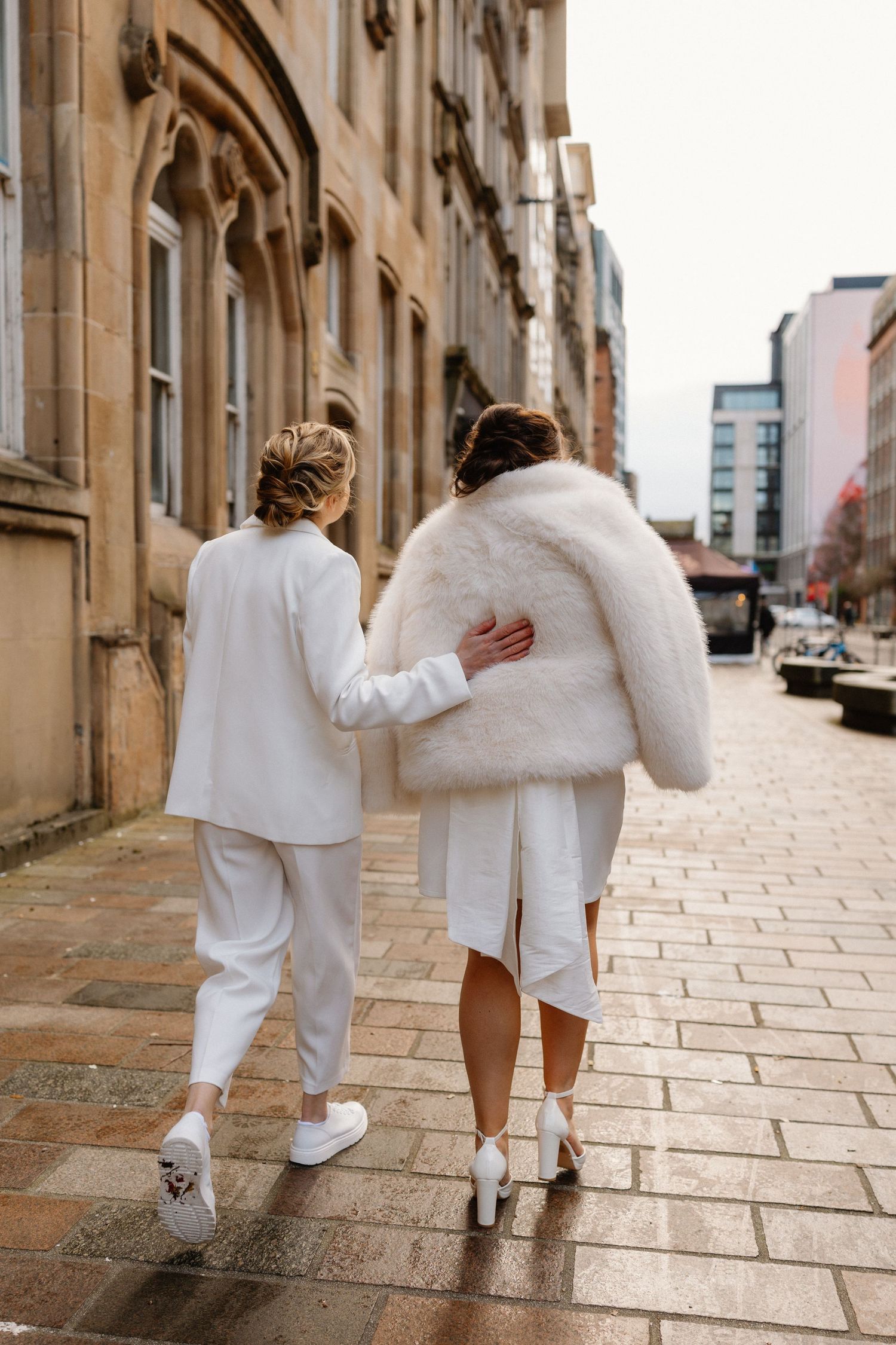 Two brides walk arm in arm down a cobblestone street, one in a white suit and one in a fur coat.