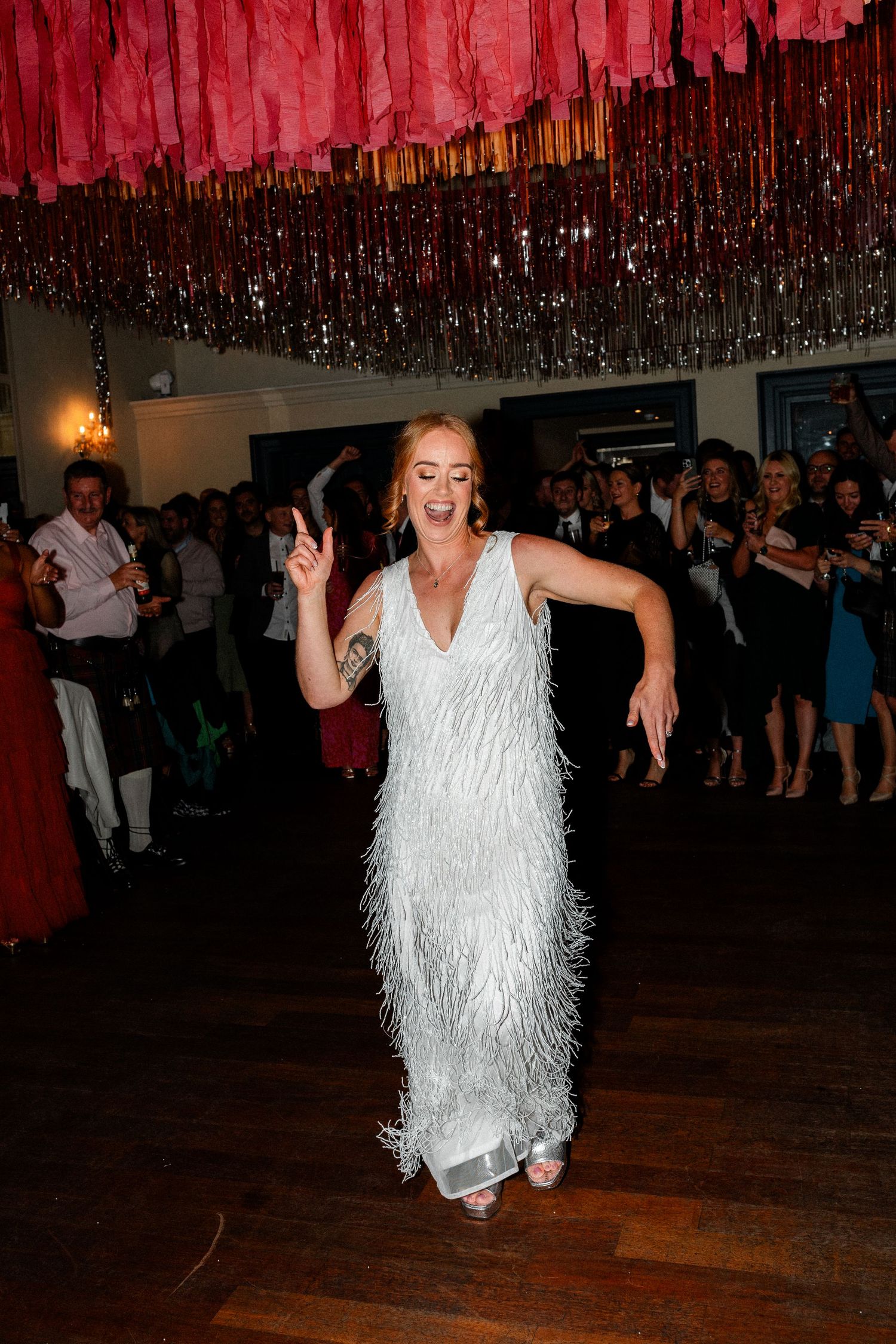 Bride dancing joyfully in a white fringed dress under pink and silver streamers at wedding reception.