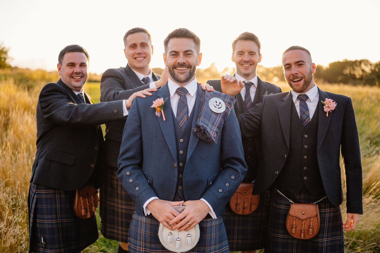 Groom and groomsmen in Scottish kilts posing outdoors at golden hour for a traditional wedding photo.