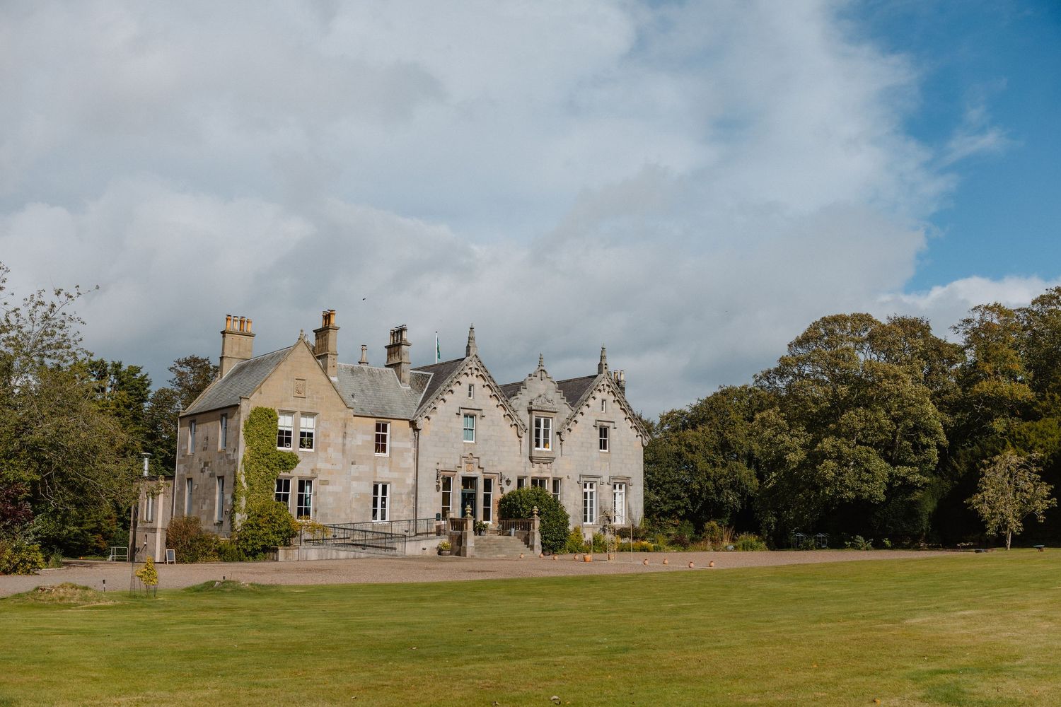A grand Scottish country manor house with stone facade set amid lush green lawns and mature trees under a blue sky.