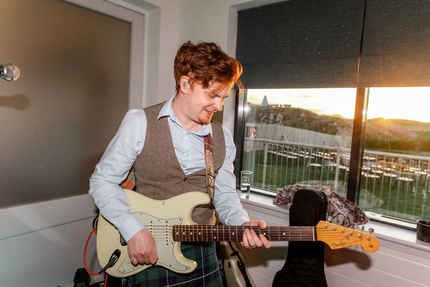 Young man playing a Fender electric guitar by a window at golden sunset hour.