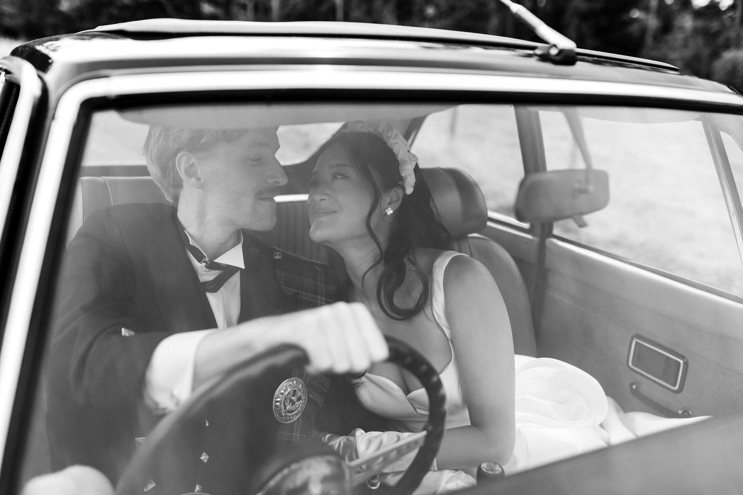 Black and white photo of bride and groom sharing romantic moment inside vintage car on wedding day.