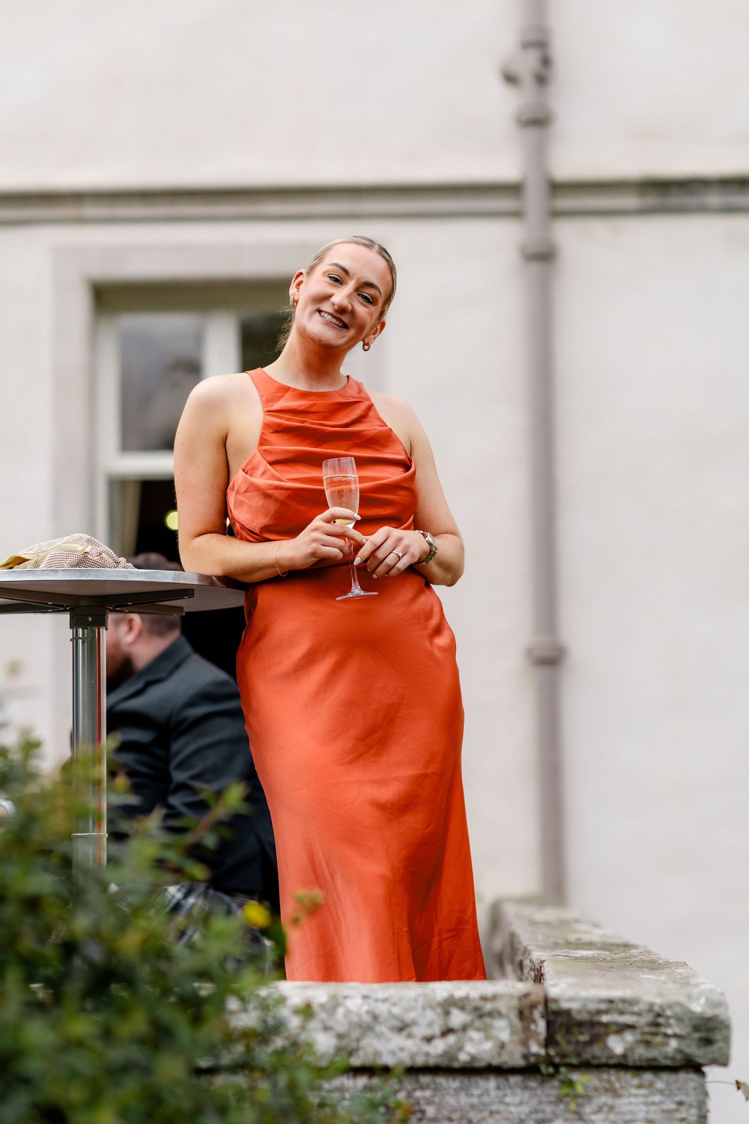 Elegant wedding guest in orange satin halter dress holding champagne flute, smiling outdoors at Scottish country house.
