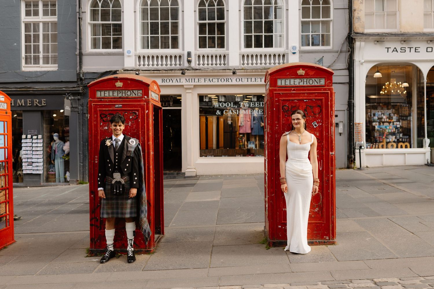 Bride and groom posing in red telephone boxes on a cobblestone street in Edinburgh, Scotland.