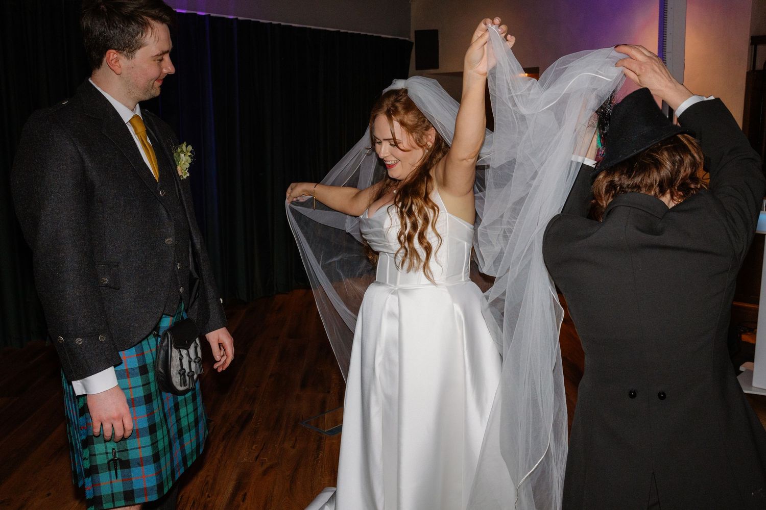 Bride in white wedding gown and veil being helped by guests while groom in Scottish kilt looks on.