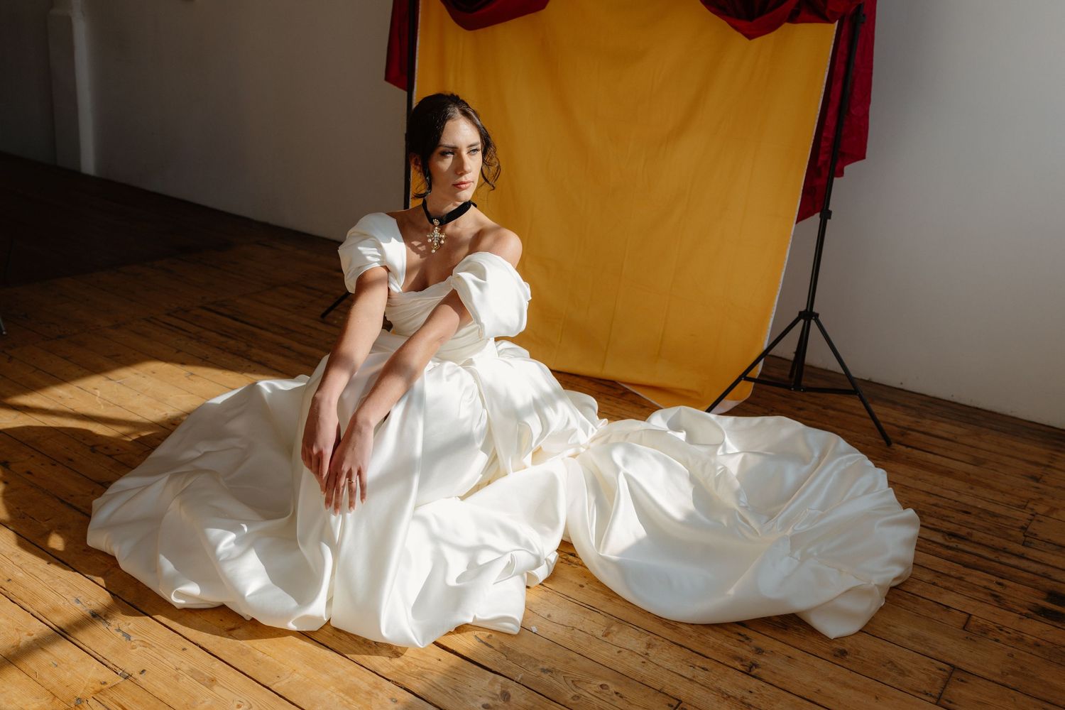 Woman in elegant white off-shoulder ballgown sitting on wooden floor in photography studio with yellow backdrop.